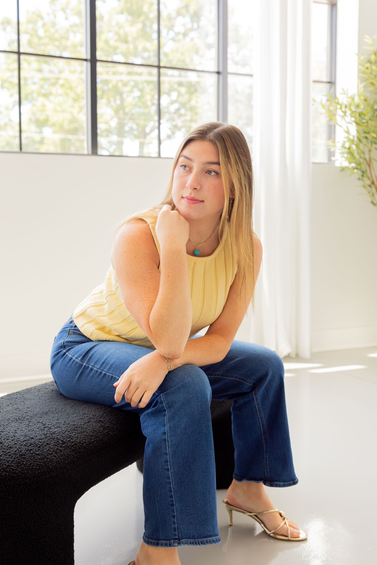 Young woman in yellow sleeveless top and wide-leg jeans sitting on a black bench near a bright window.
