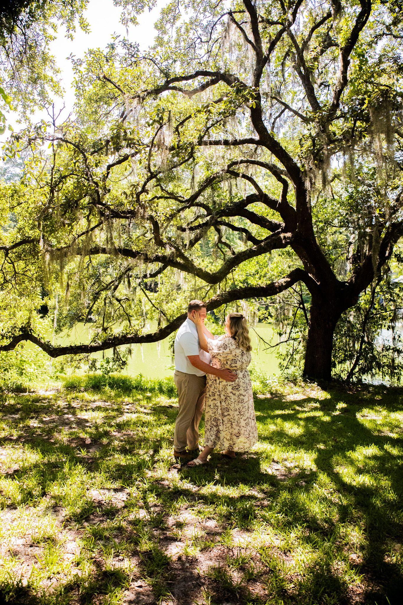 A couple embraces under a sprawling oak tree draped in Spanish moss in a lush green park setting.