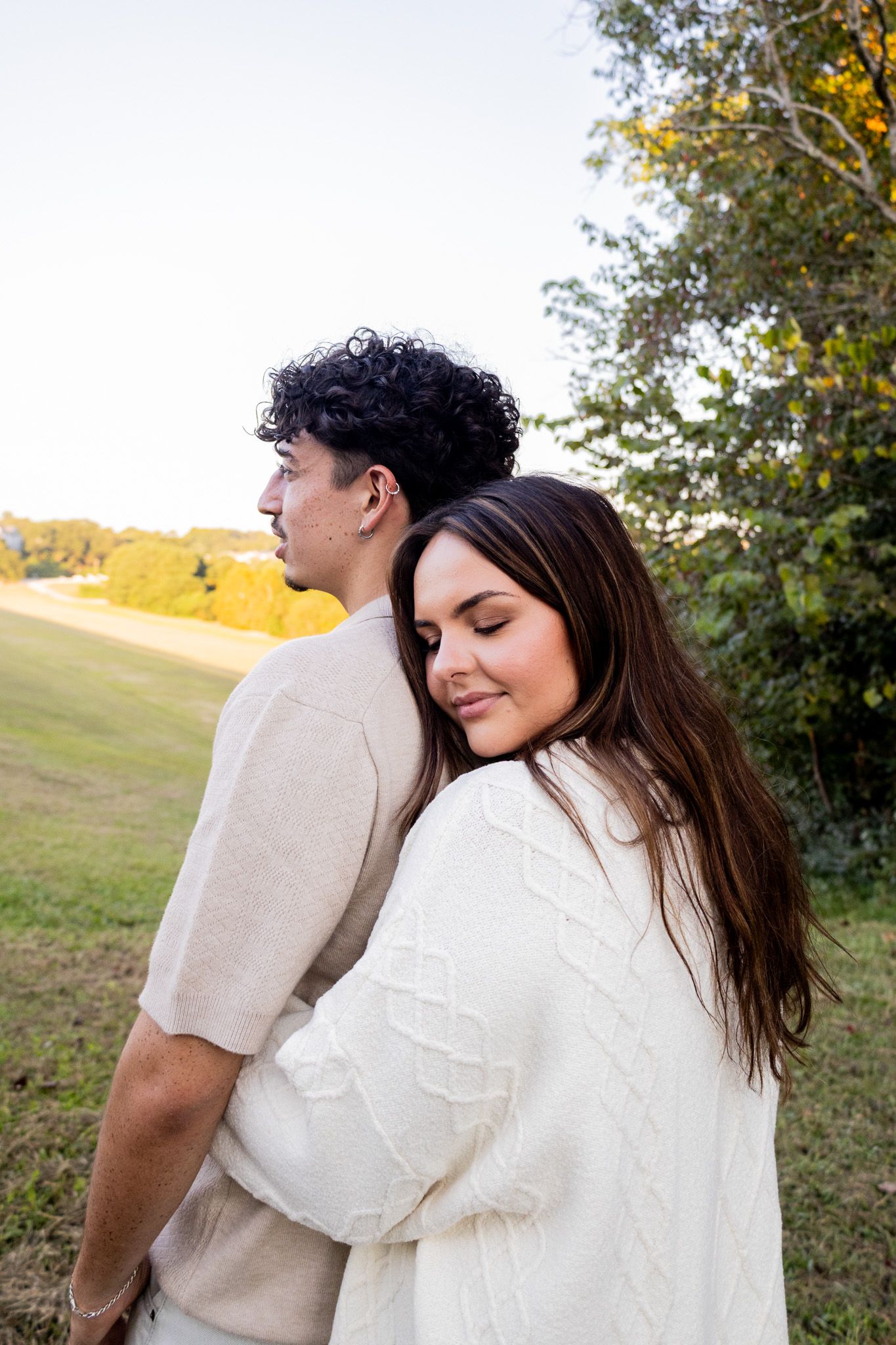 A woman in a white cable-knit sweater rests her head on a man's shoulder outdoors in a golden autumn setting.