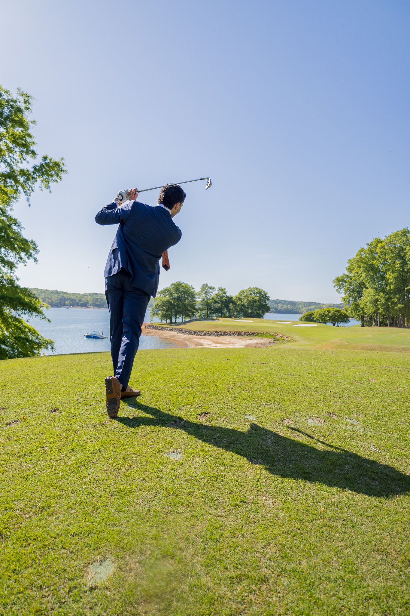 Man in suit swinging golf club on lush green course with lake and trees in background on sunny day.