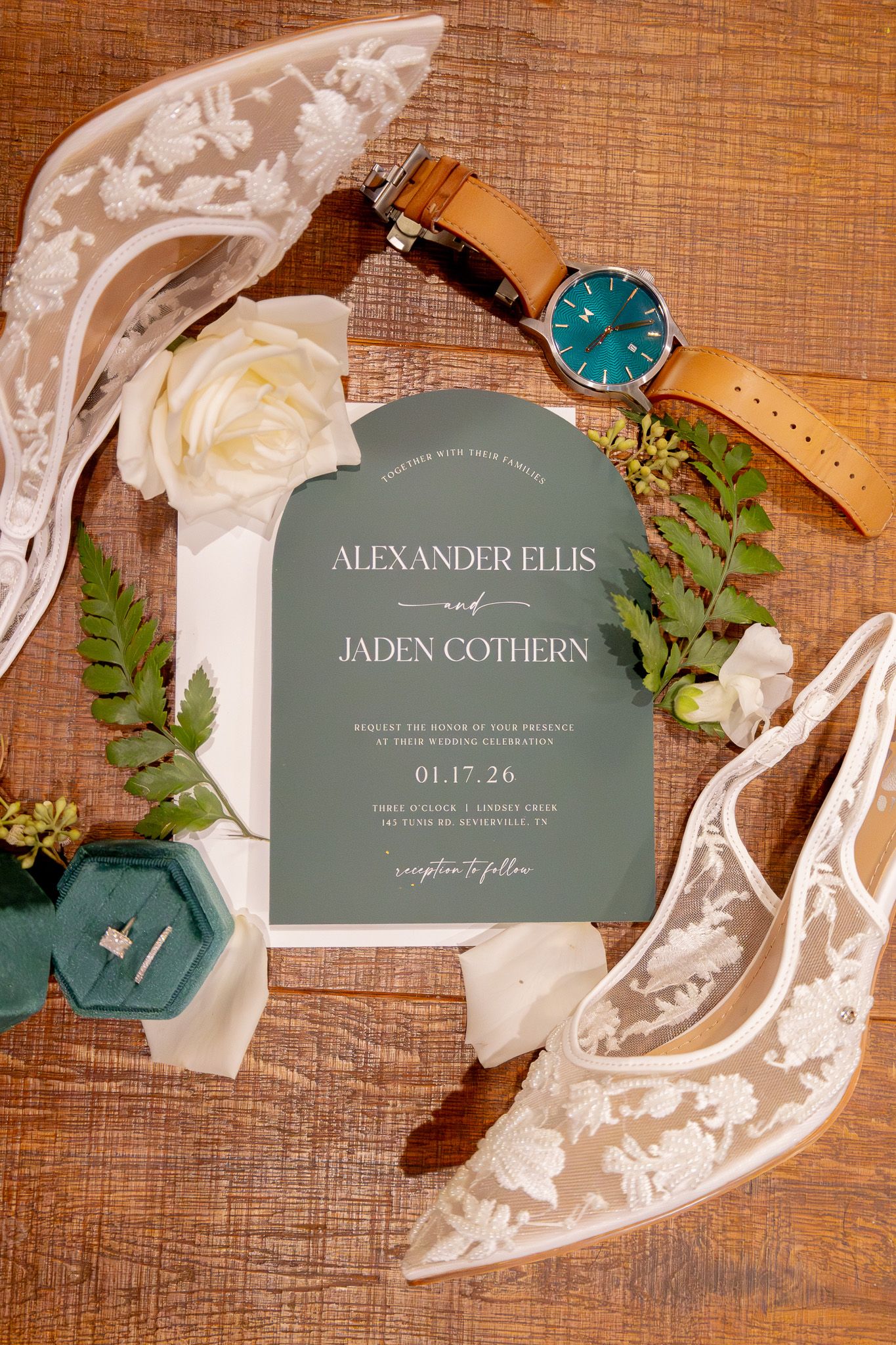 Elegant wedding invitation for Alexander Ellis and Jaden Cothern styled with lace heels, a watch, white roses, and greenery on wood.