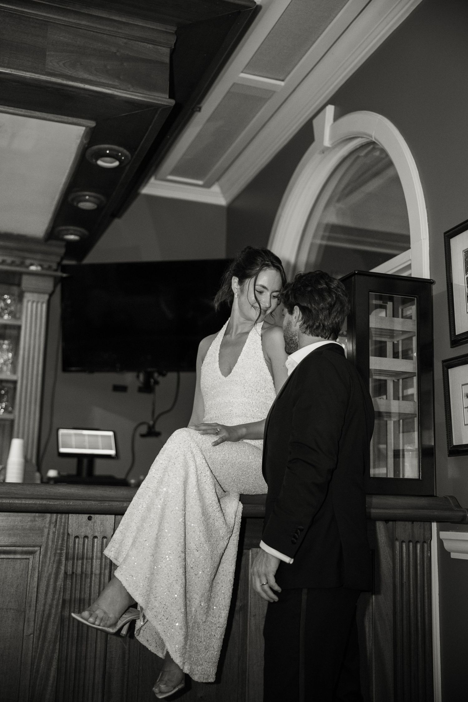 Black and white photo of a couple sharing an intimate moment at a bar, bride in white dress seated on counter.