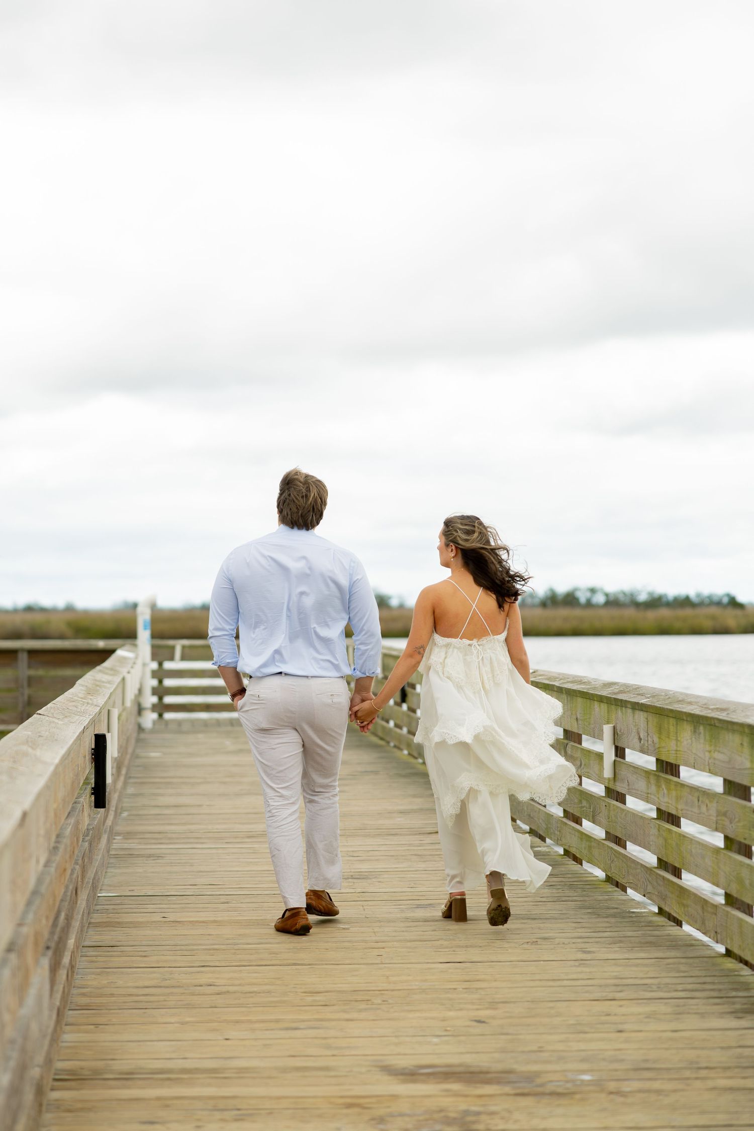 Couple holding hands and walking away on a wooden pier overlooking calm water under a cloudy sky, the woman in a flowing white dress.