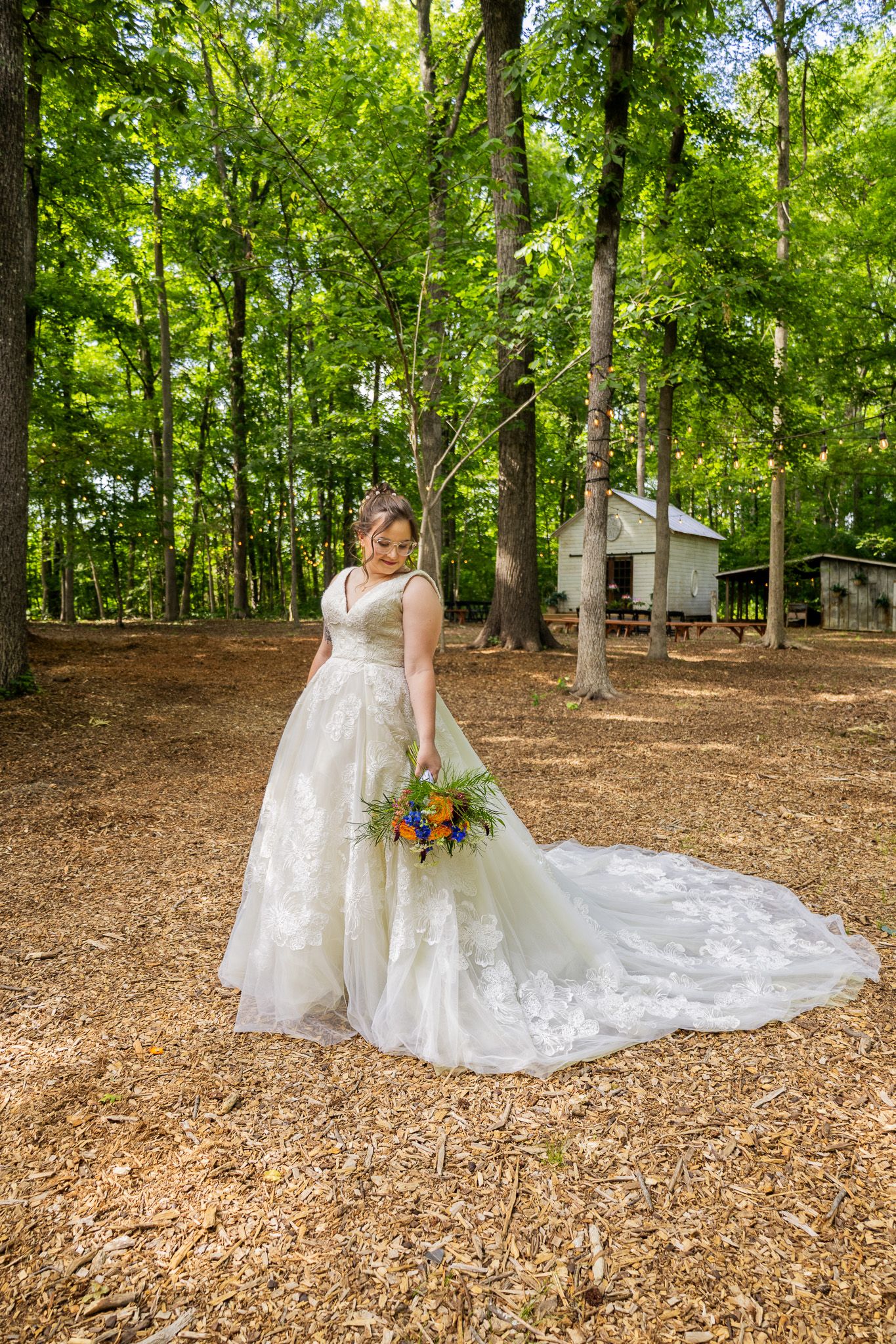 Bride in white lace gown holding colorful bouquet stands in wooded outdoor venue with wood chip ground.