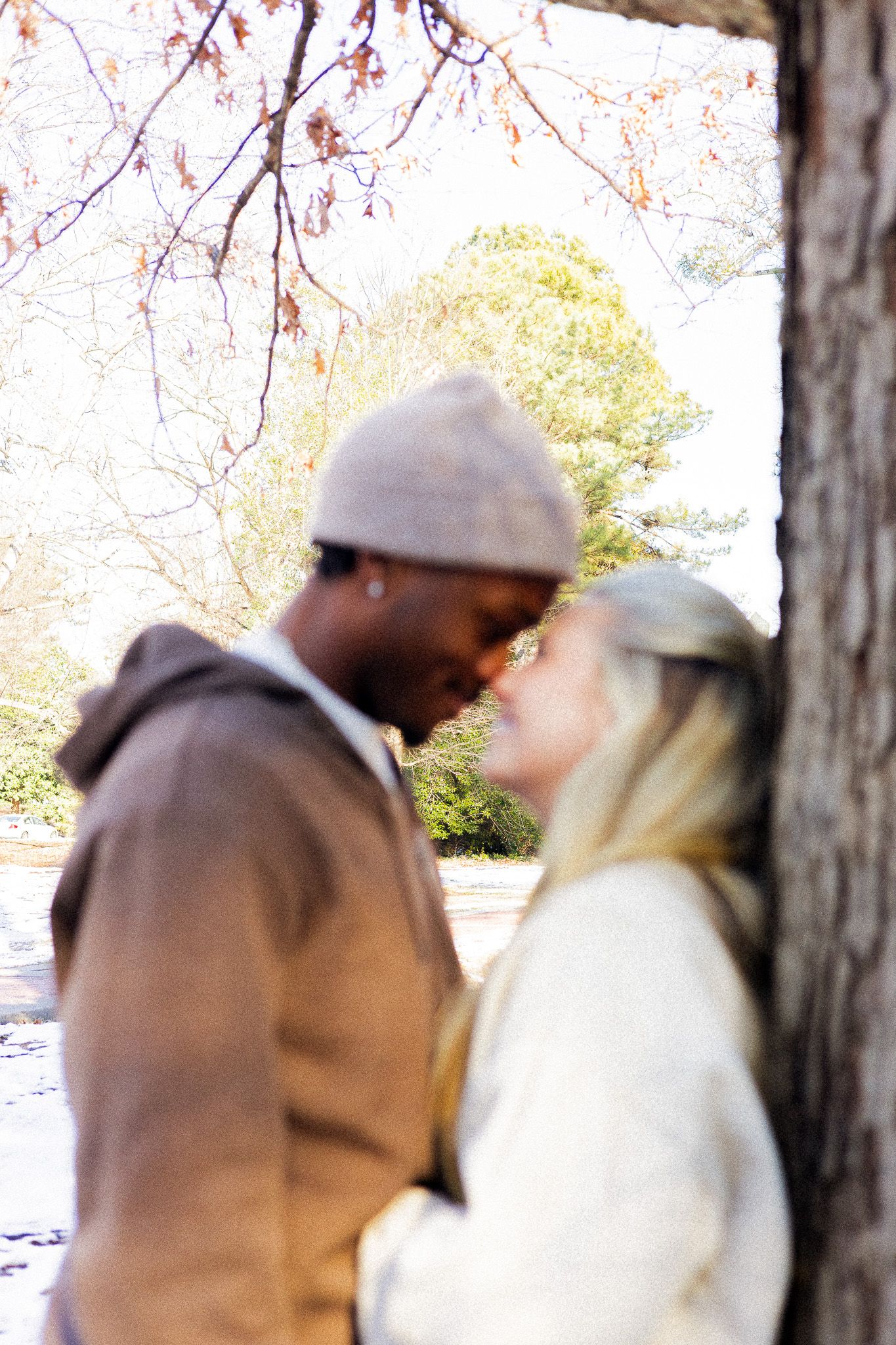 Interracial couple sharing a romantic moment outdoors in autumn, framed by tree trunk and colorful fall foliage.