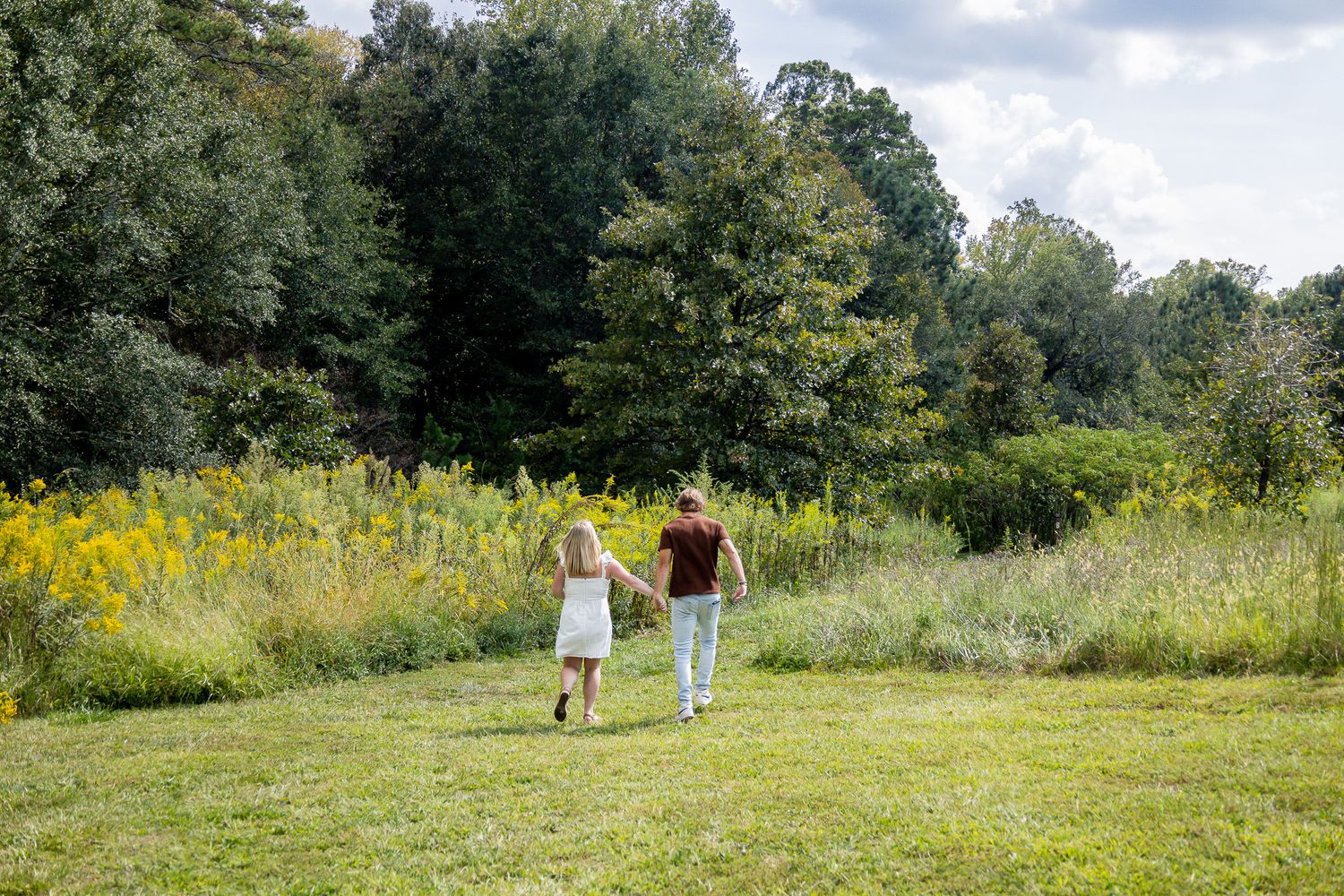 Couple walking hand in hand through a lush green meadow with wildflowers and tall trees in background.