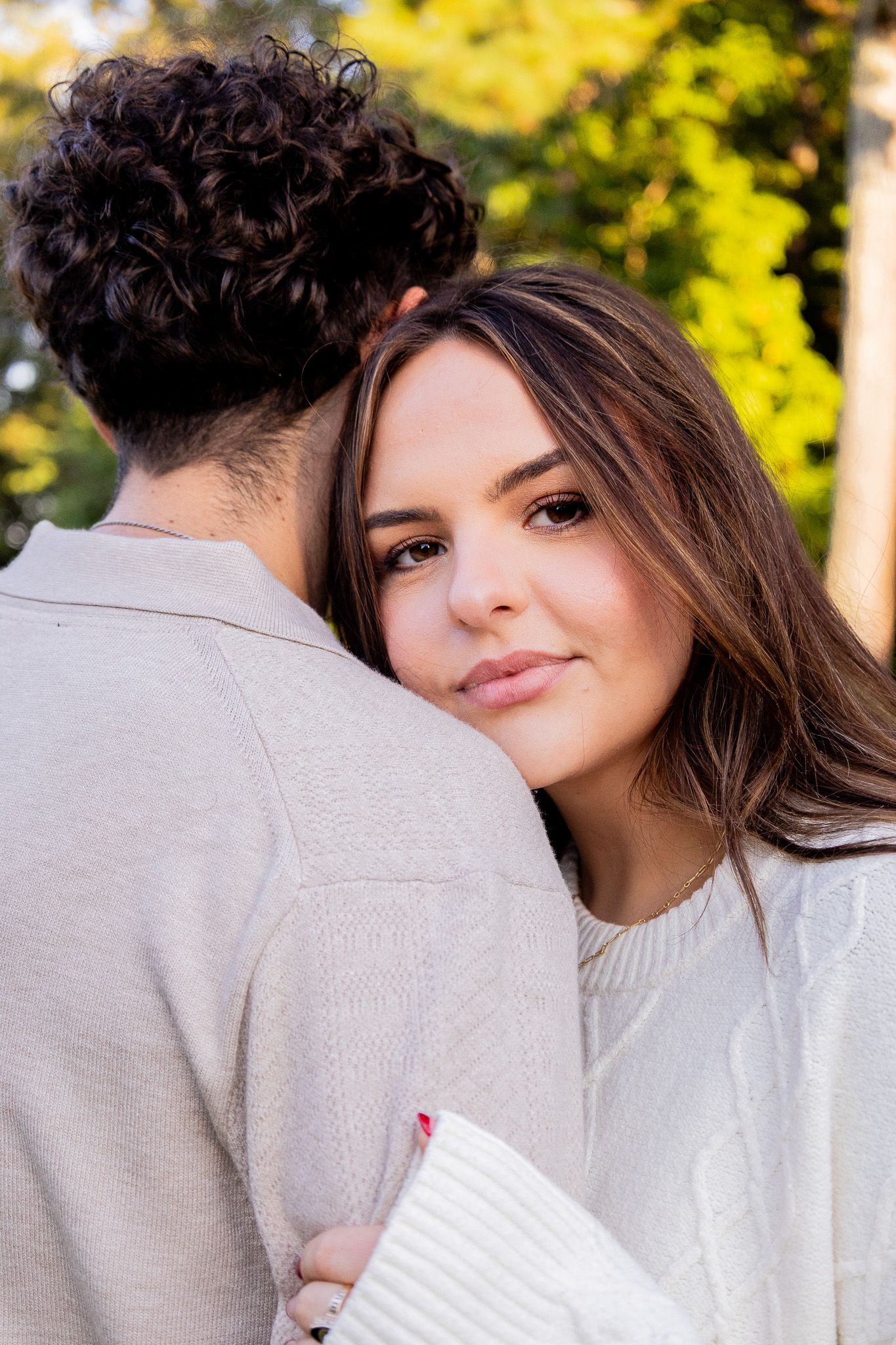 A young woman rests her head on a man's shoulder, smiling softly, while embracing outdoors in a lush green setting.