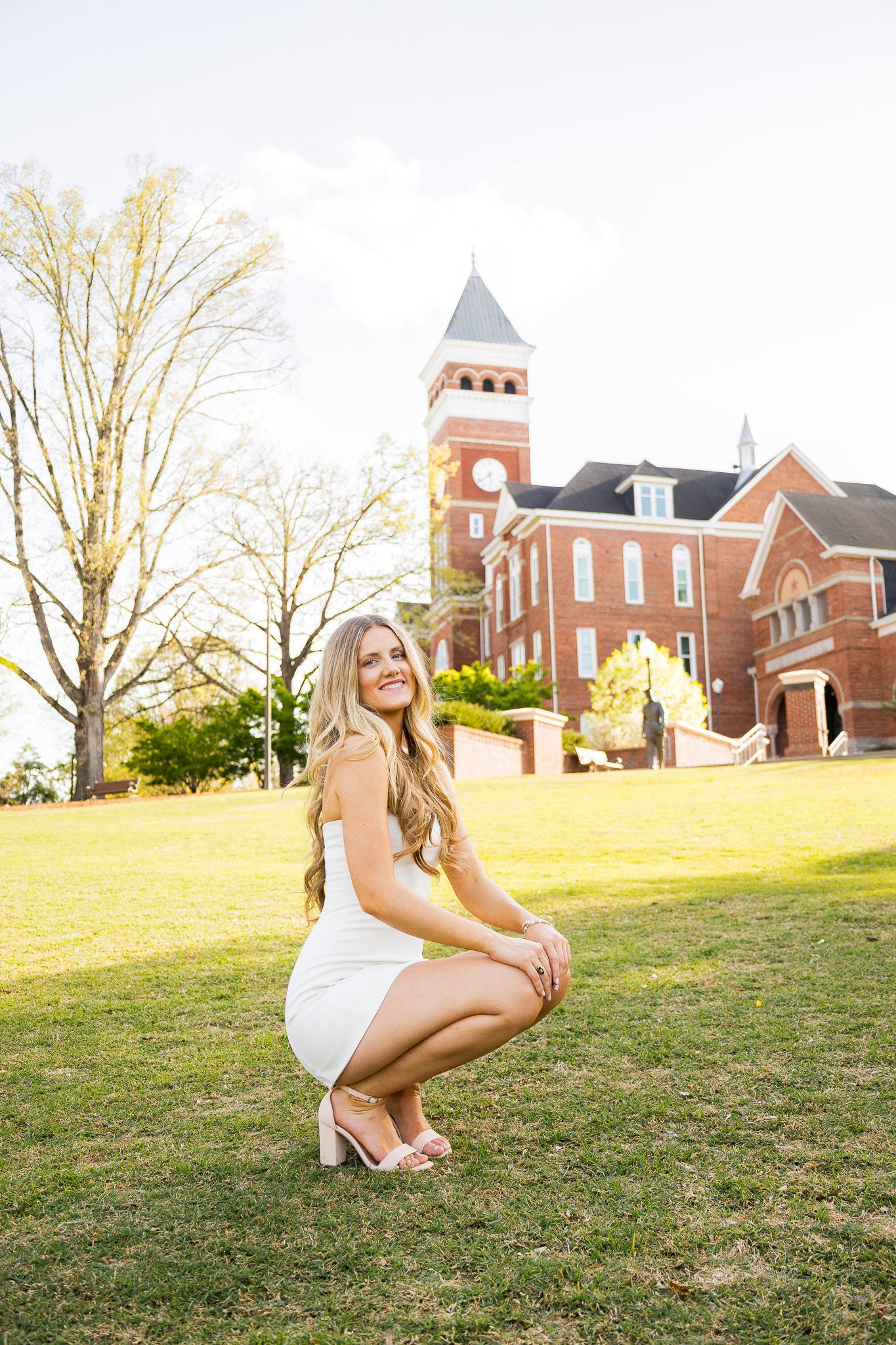Young woman in white dress crouching on green lawn in front of a red brick building with clock tower.