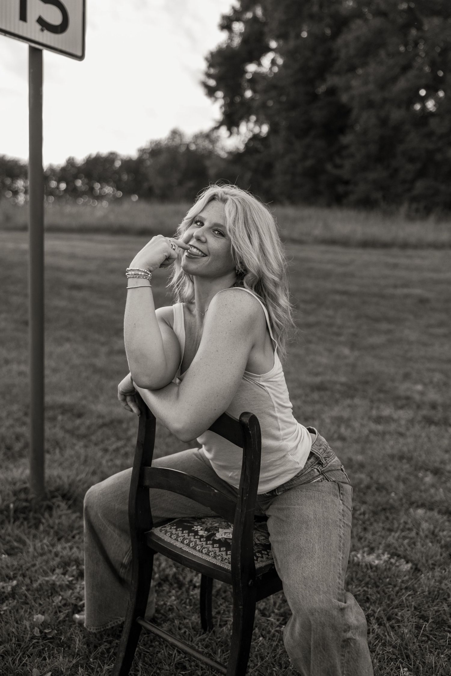 Black and white photo of a smiling blonde woman sitting on a chair outdoors near a speed limit sign.