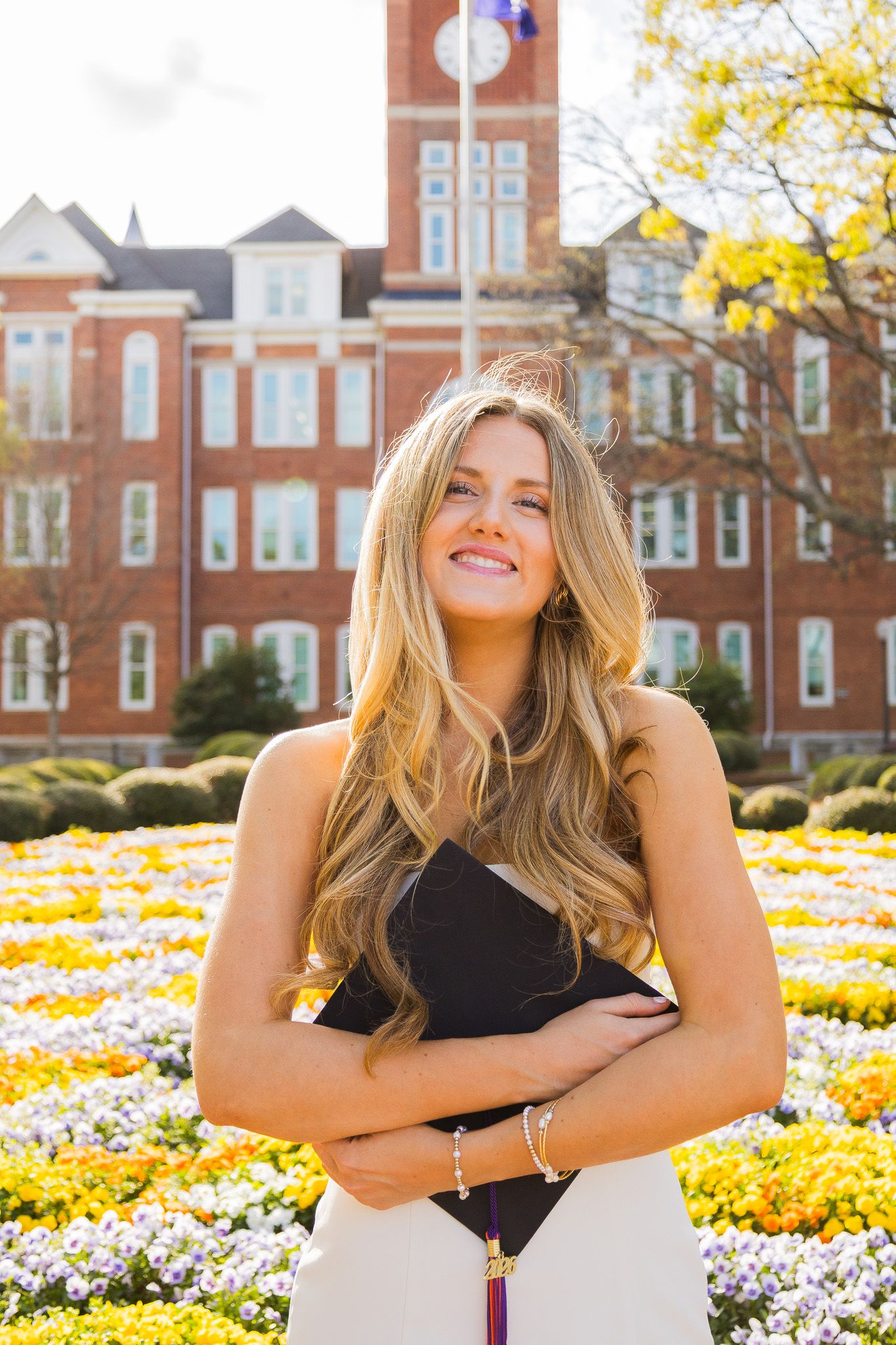Smiling blonde woman holding graduation cap in front of colorful flower garden and brick university building.