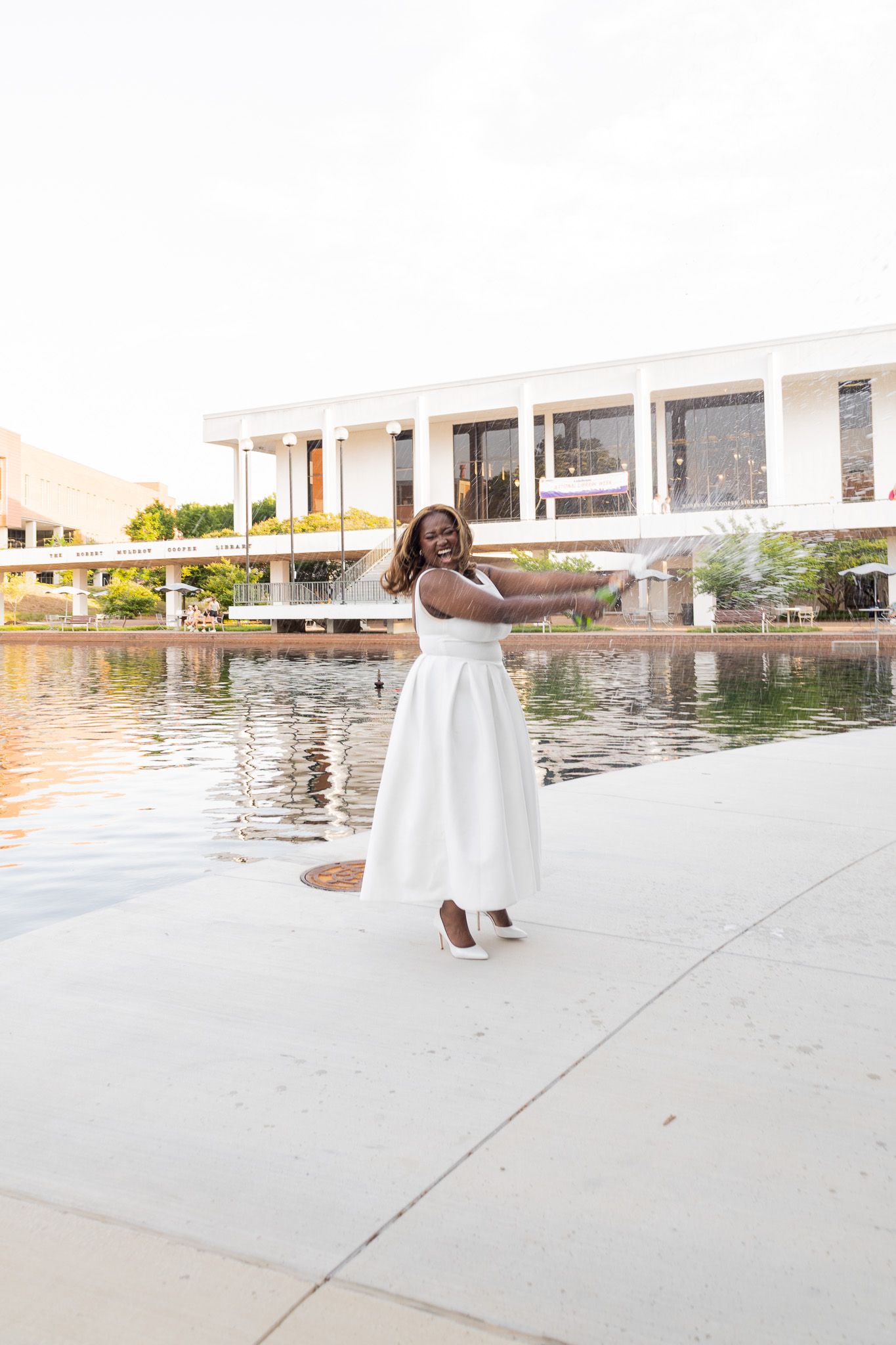 A joyful Black woman in a white strapless dress poses by a reflective pool near a modern building.