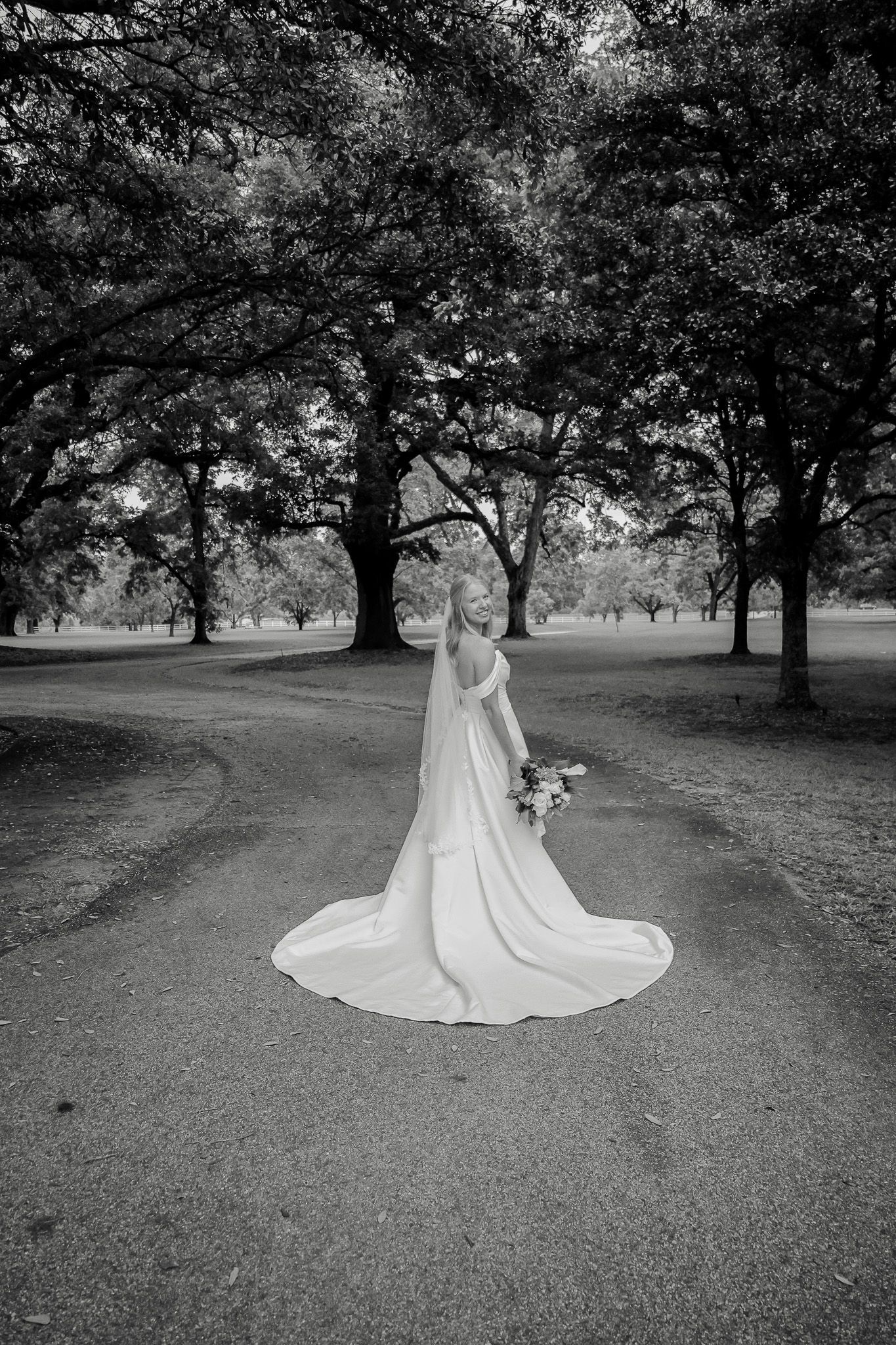 Black and white photo of a bride in a flowing gown and veil holding a bouquet, standing on a path surrounded by trees.