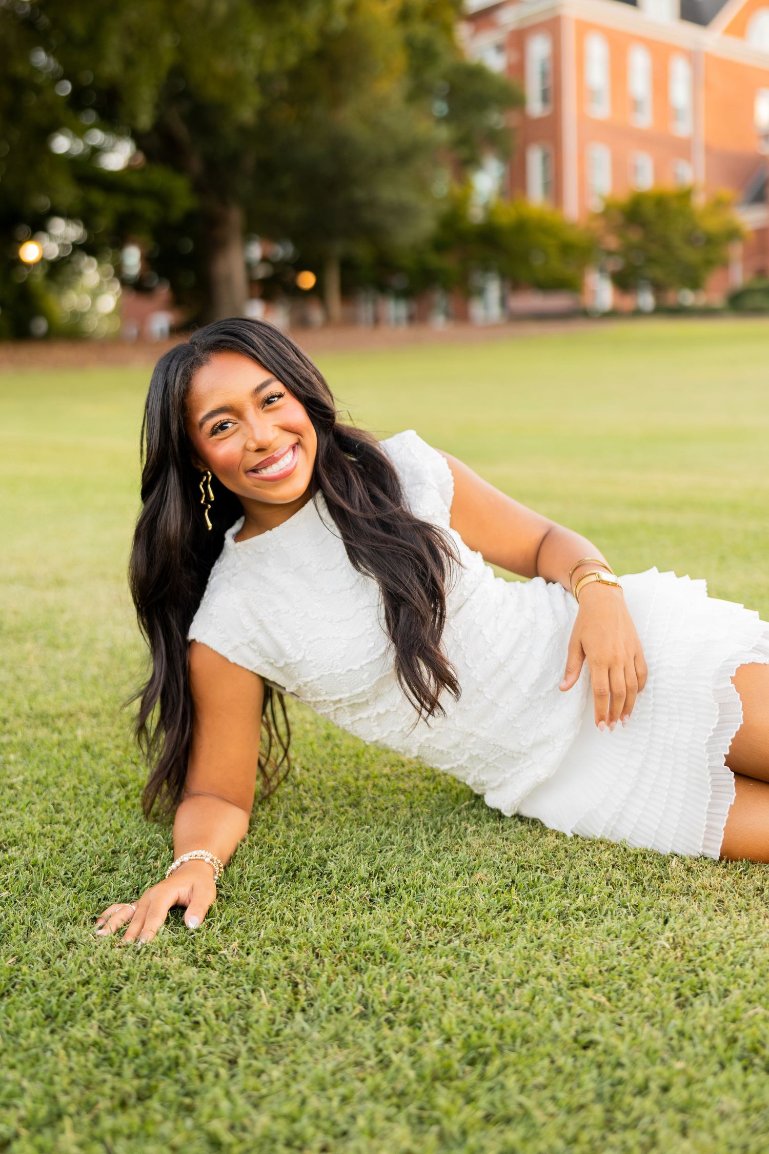 Young woman in white dress poses lying on green grass lawn with brick building in background.