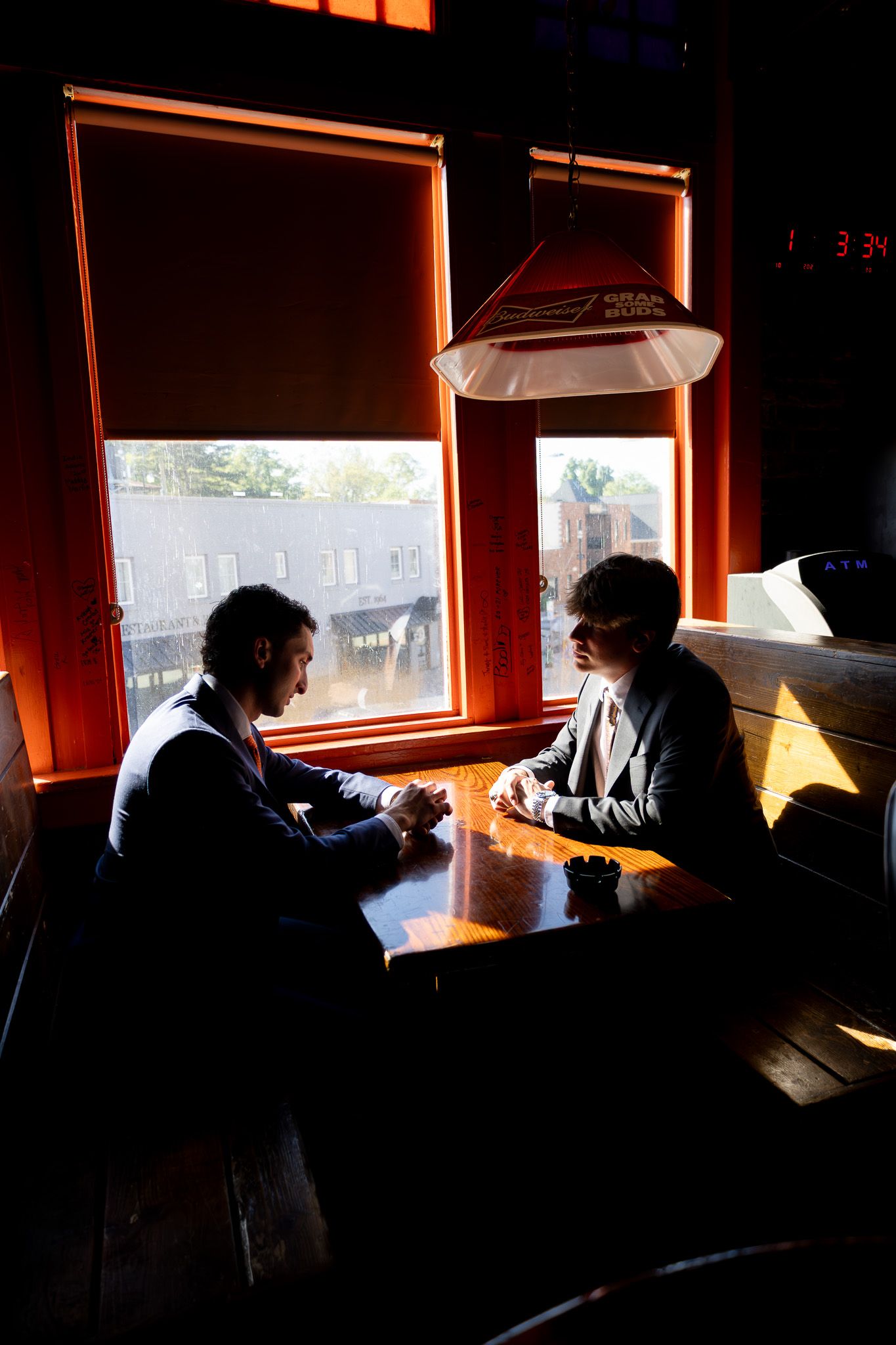 Two businessmen sit across from each other at a wooden table in a dimly lit restaurant booth near sunlit windows.