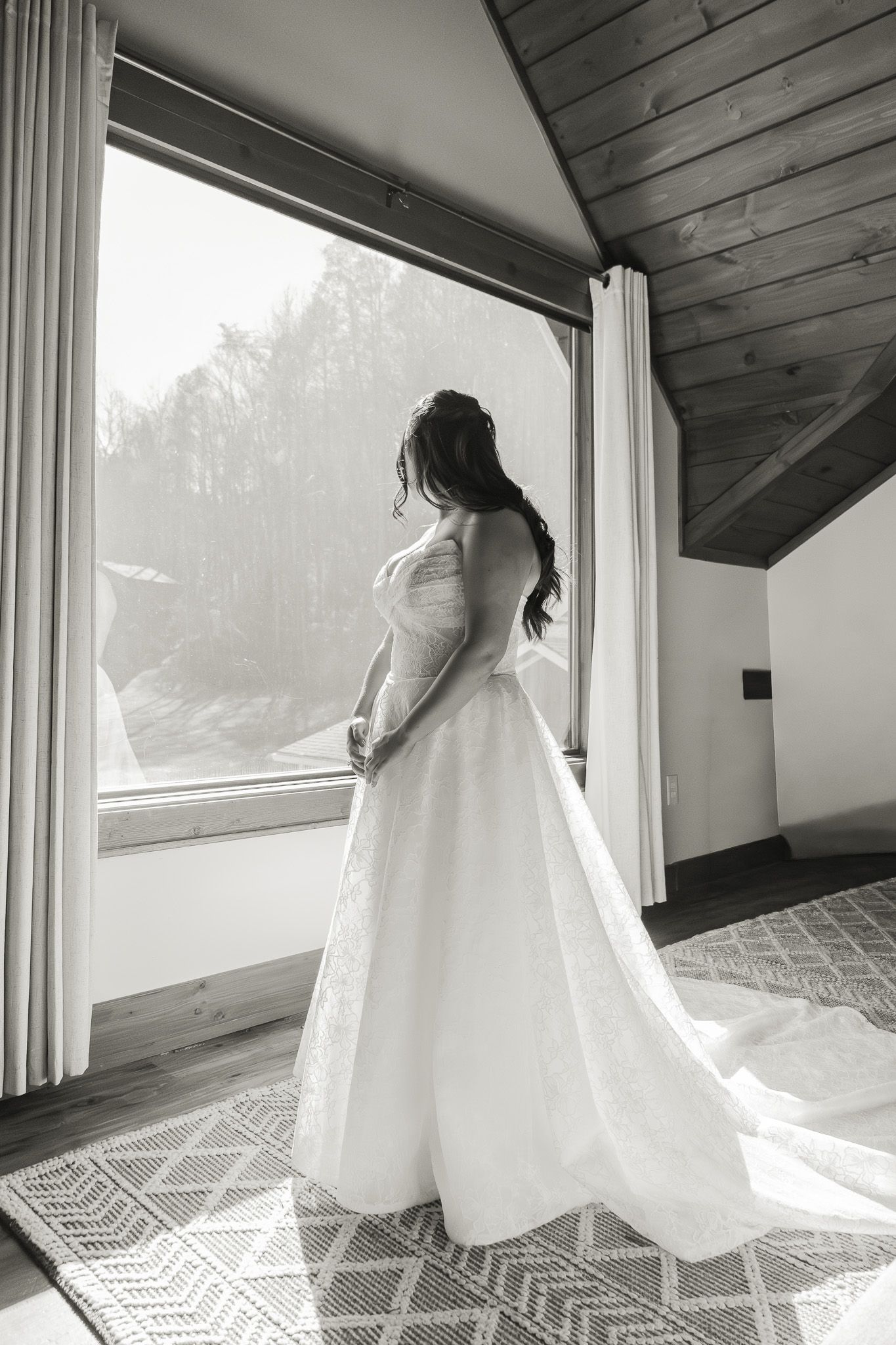 Bride in lace wedding gown stands before large window gazing outside in black and white bridal portrait.