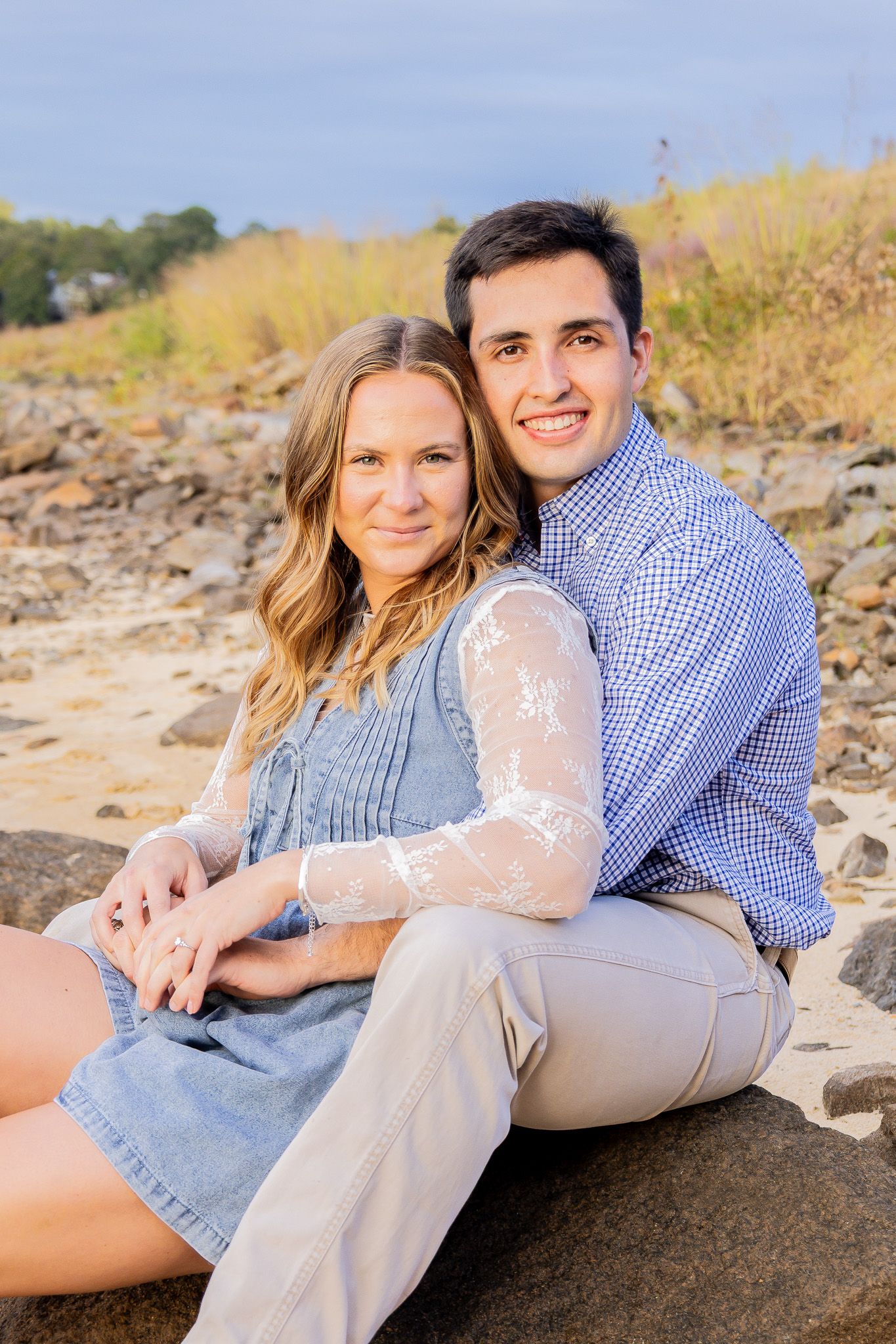 A smiling couple sits on rocks outdoors, woman in denim dress, man in blue plaid shirt, posing for engagement photo.