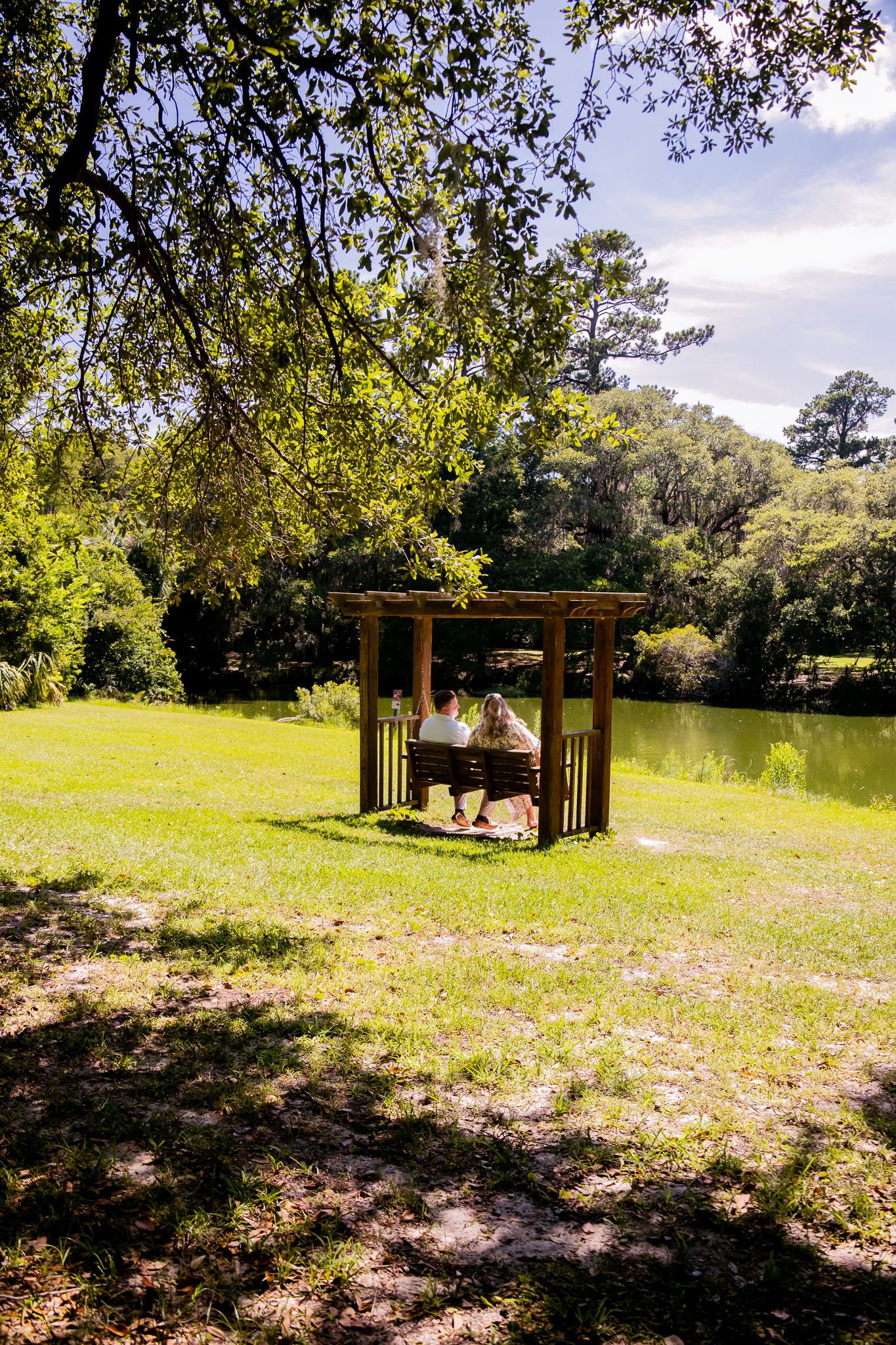 Couple sitting on a wooden pergola swing bench by a serene pond surrounded by lush green trees in a park.