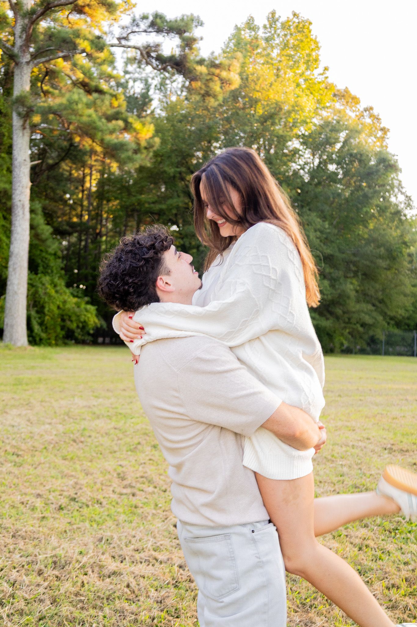 A man lifts a woman in a warm embrace outdoors in a sunny park surrounded by lush green trees.