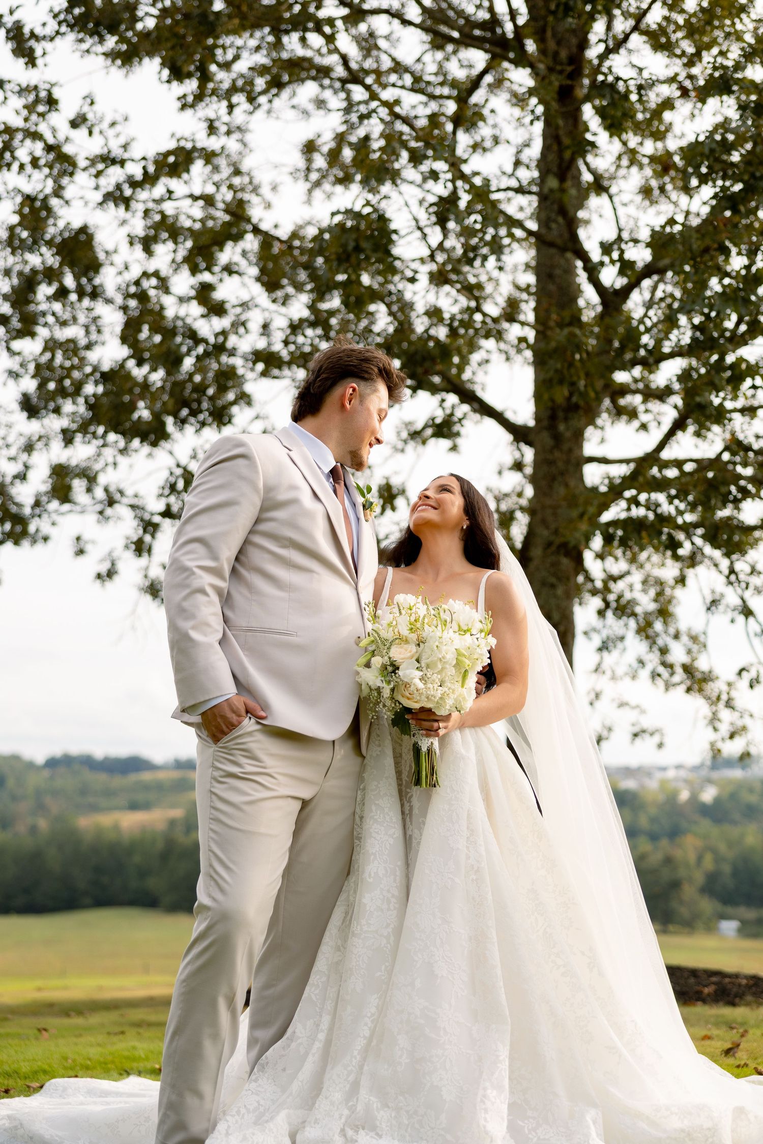 A bride and groom share a joyful moment outdoors under a large tree, she holds a white bouquet in a lace gown.