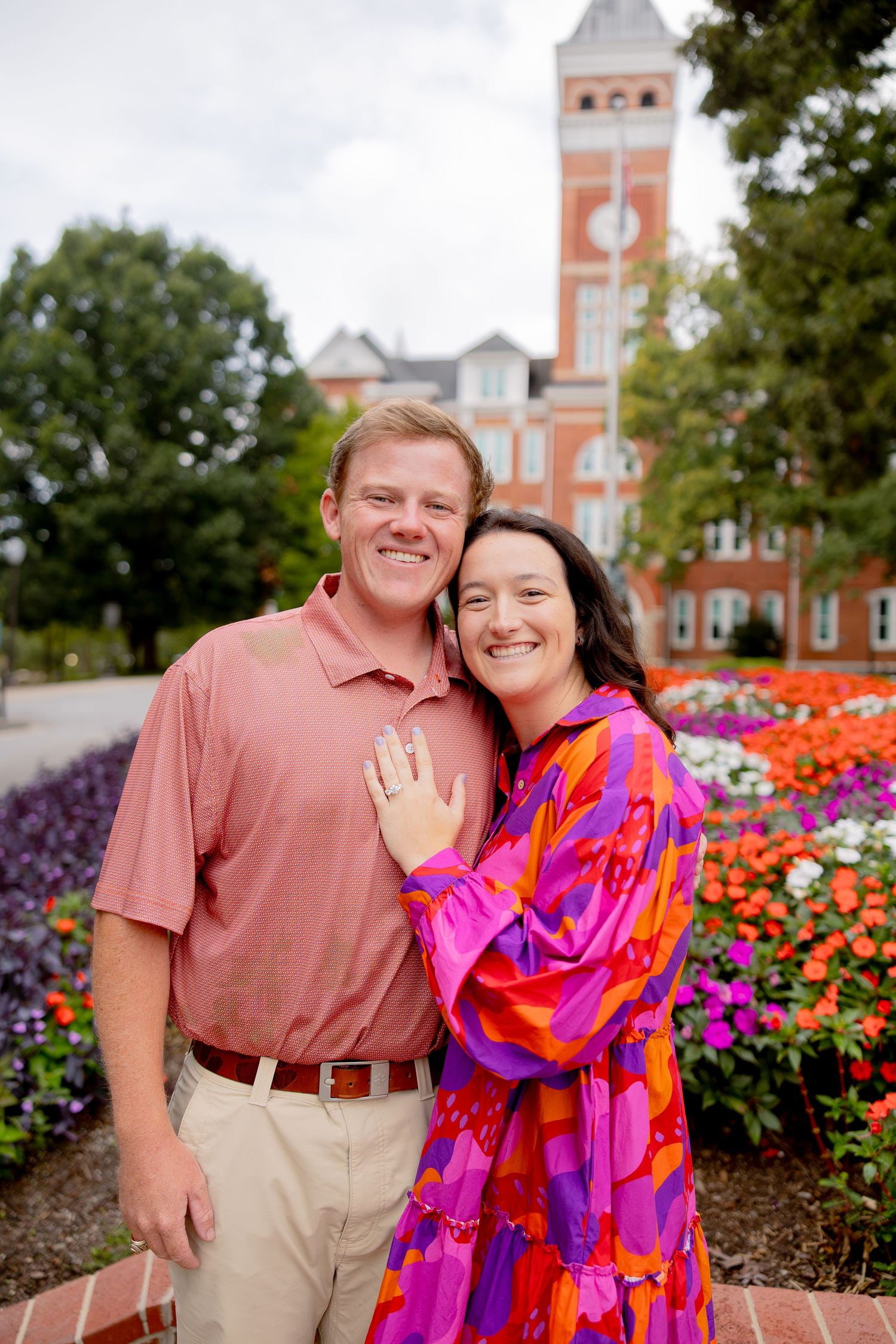 Happy couple posing in front of a brick clock tower building surrounded by colorful flower gardens.