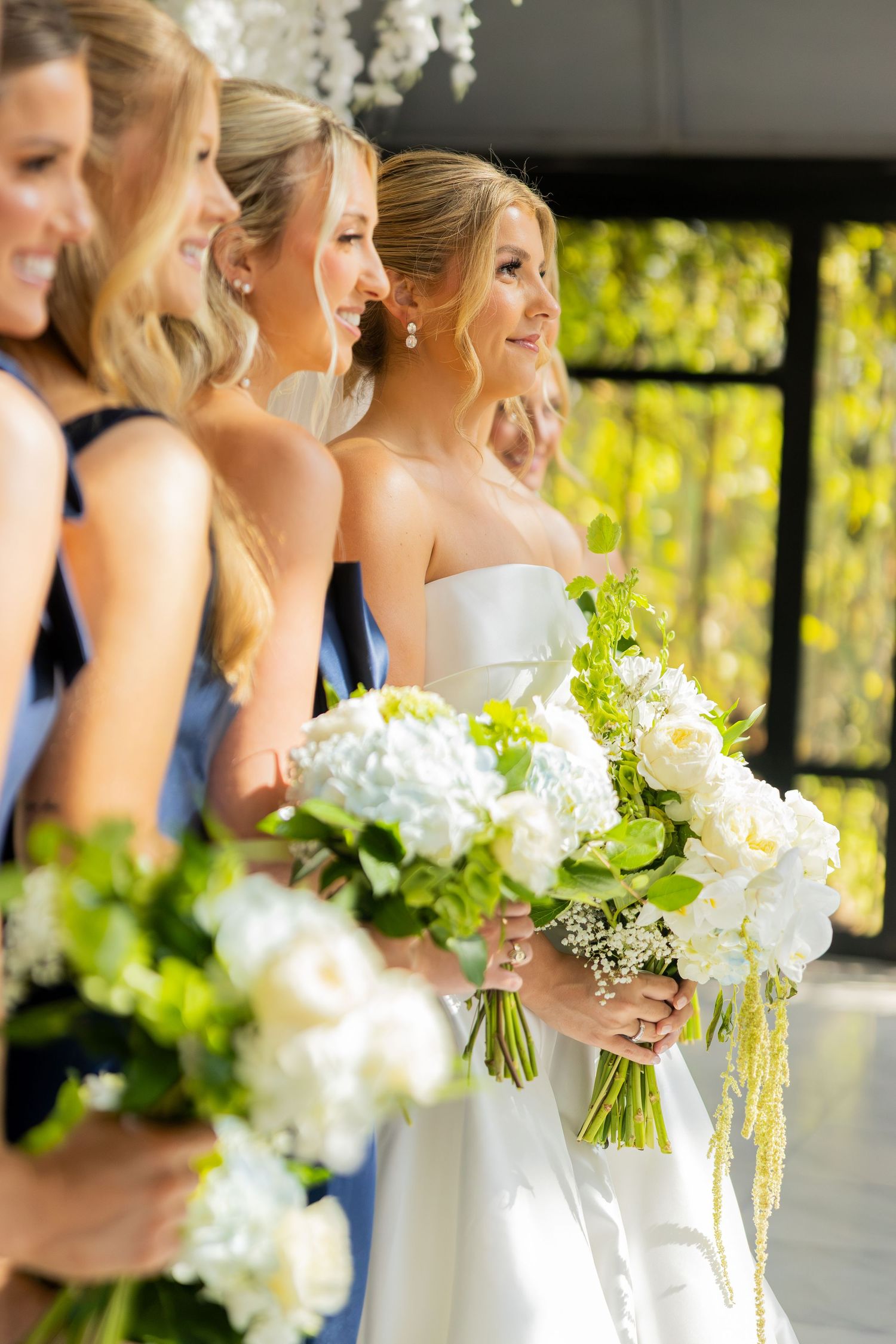 Bride in white strapless gown holds lush white floral bouquet alongside bridesmaids in blue dresses at elegant wedding.