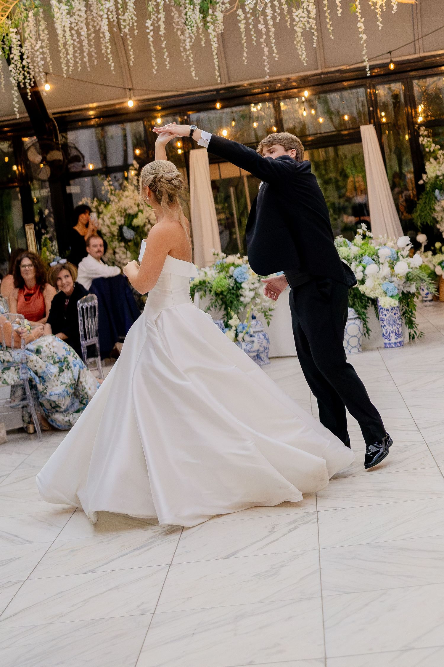Bride and groom share their first dance, twirling on a marble floor surrounded by floral arrangements and guests.