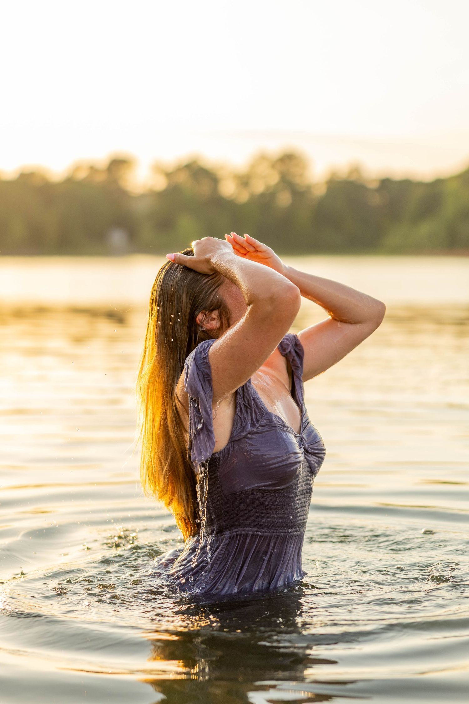 Woman standing waist-deep in golden sunset lake water, wearing wet blue dress, hands in long wet hair.