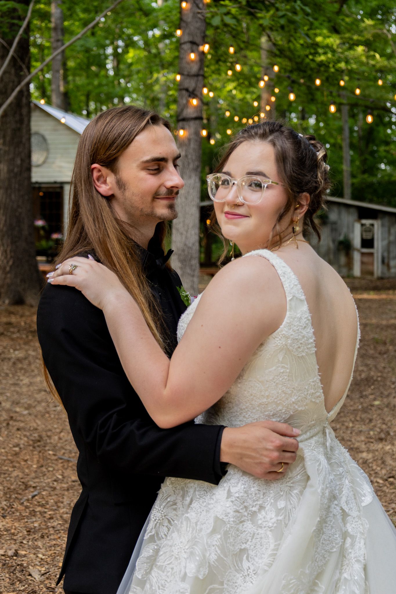 A couple in wedding attire embraces outdoors, bride in white lace gown with glasses, groom in black suit, string lights behind.