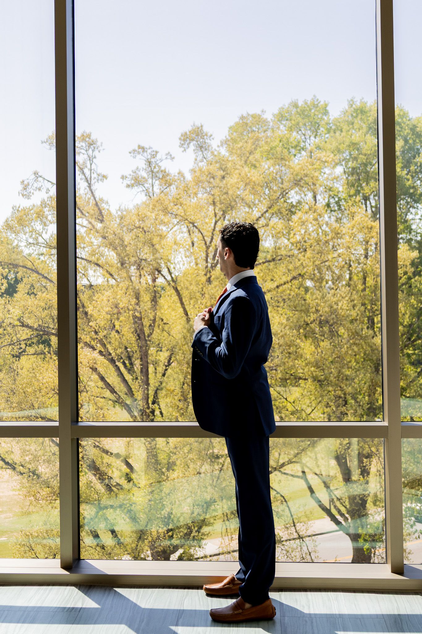 Businessman in navy suit standing by large windows overlooking autumn trees, adjusting his tie.