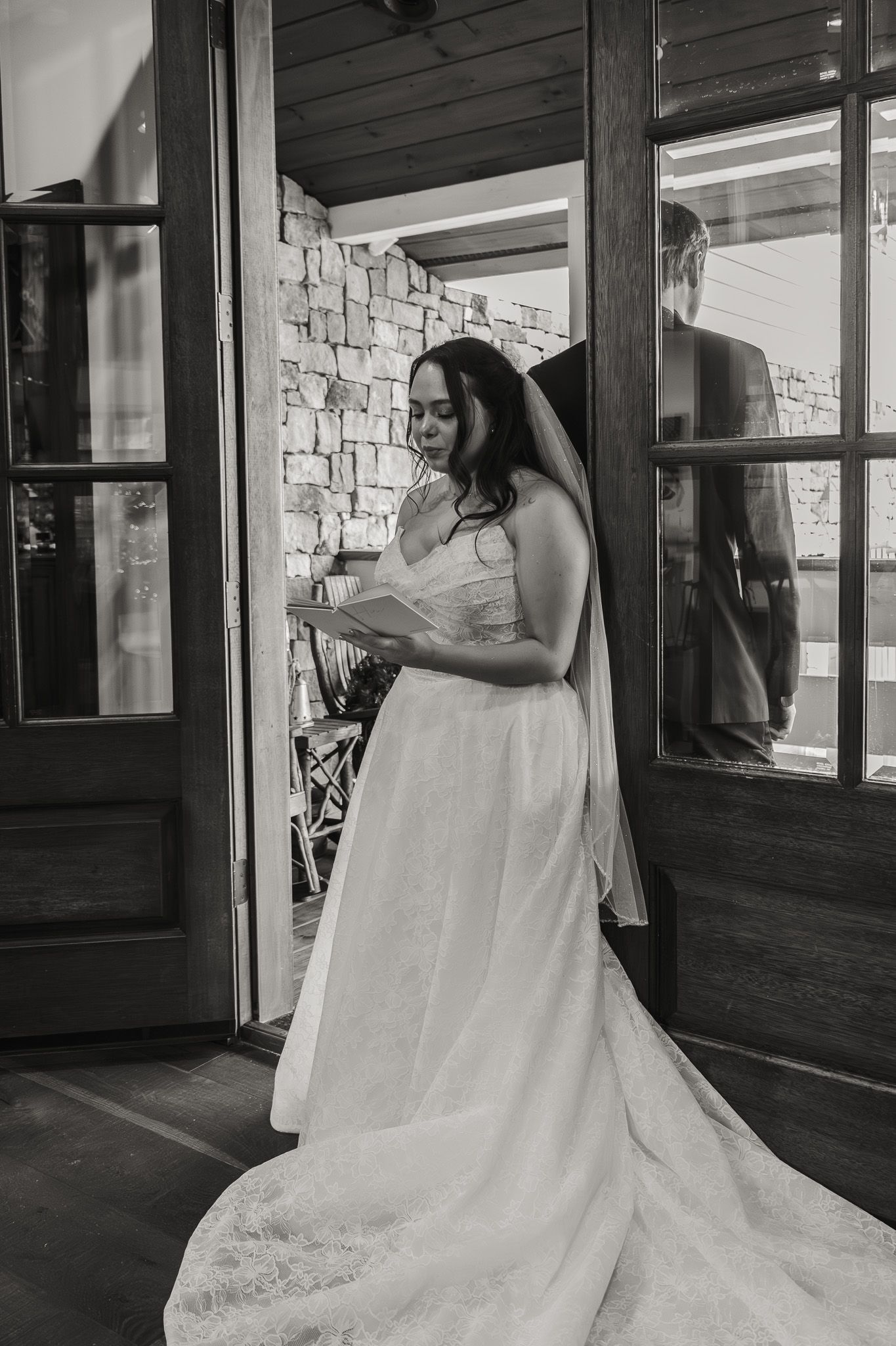 Bride in white gown and veil reads vows at rustic venue doorway in black and white wedding photo.
