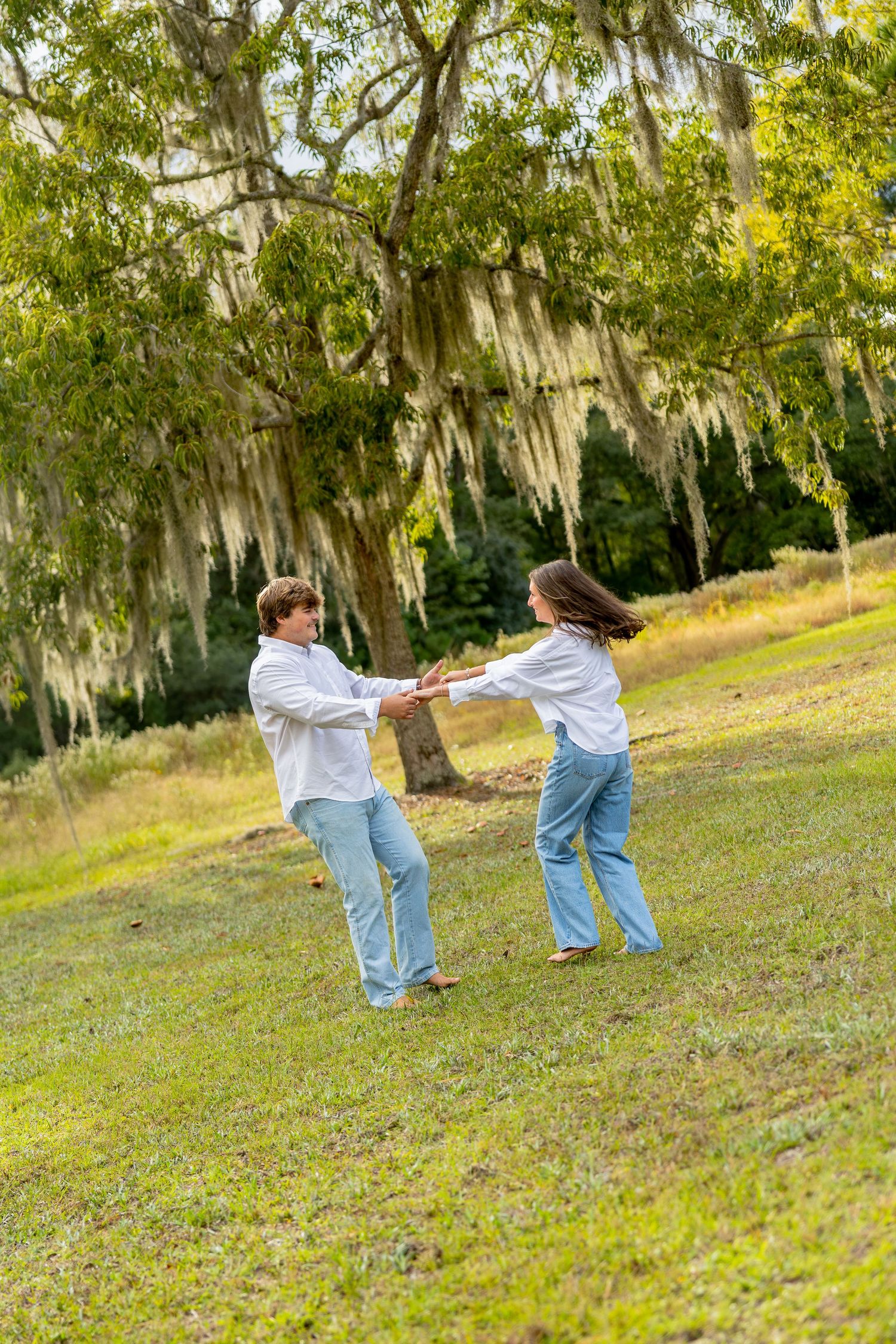 Two people in white shirts and blue jeans hold hands and spin together on a grassy lawn beneath a live oak draped in Spanish moss.