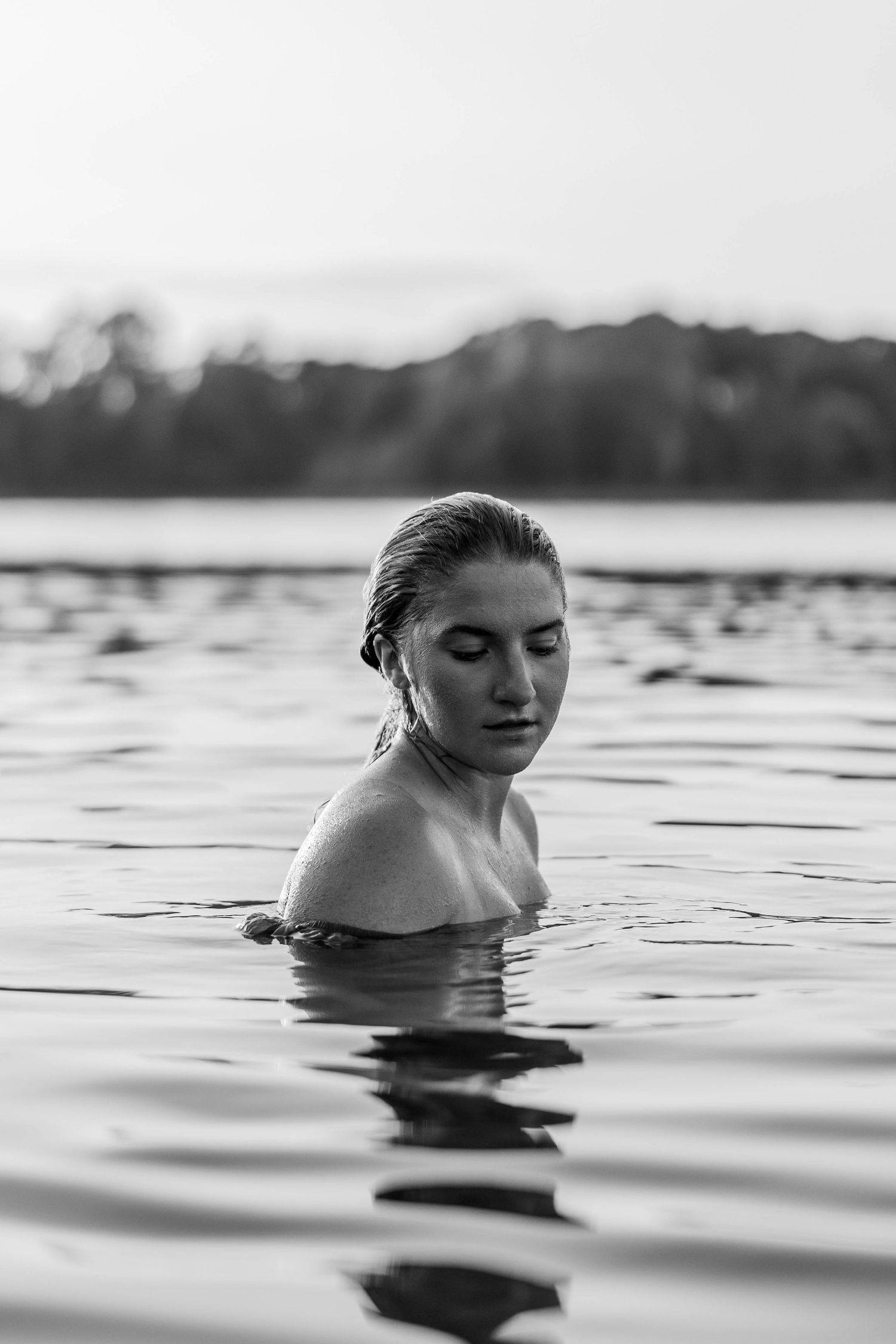 Black and white photo of a woman submerged in a calm lake, eyes closed, with trees in the background.