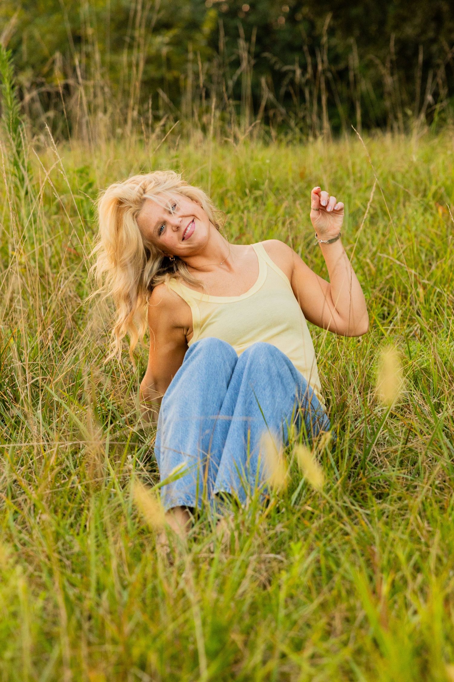 Smiling blonde woman sitting in tall green grass field wearing yellow tank top and blue jeans on a sunny day.