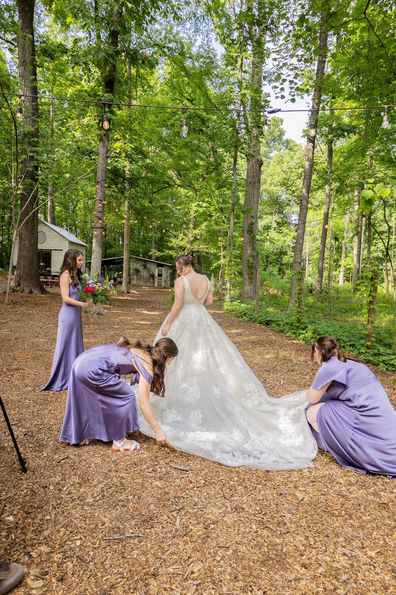Bride in white gown with bridesmaids in purple dresses adjusting her train in a wooded outdoor wedding setting.