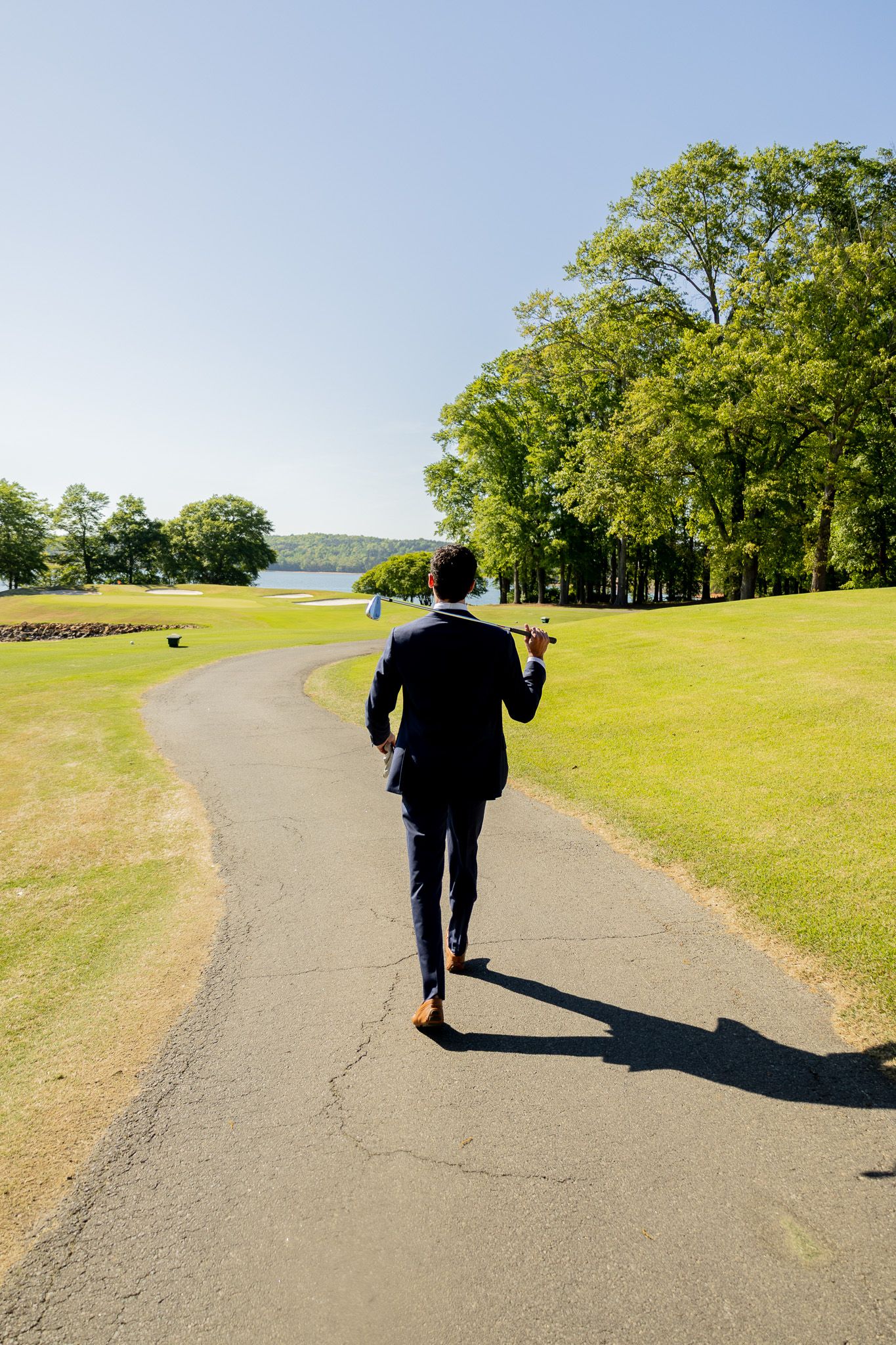 Businessman walking down golf course path carrying club with scenic lake and trees visible ahead.