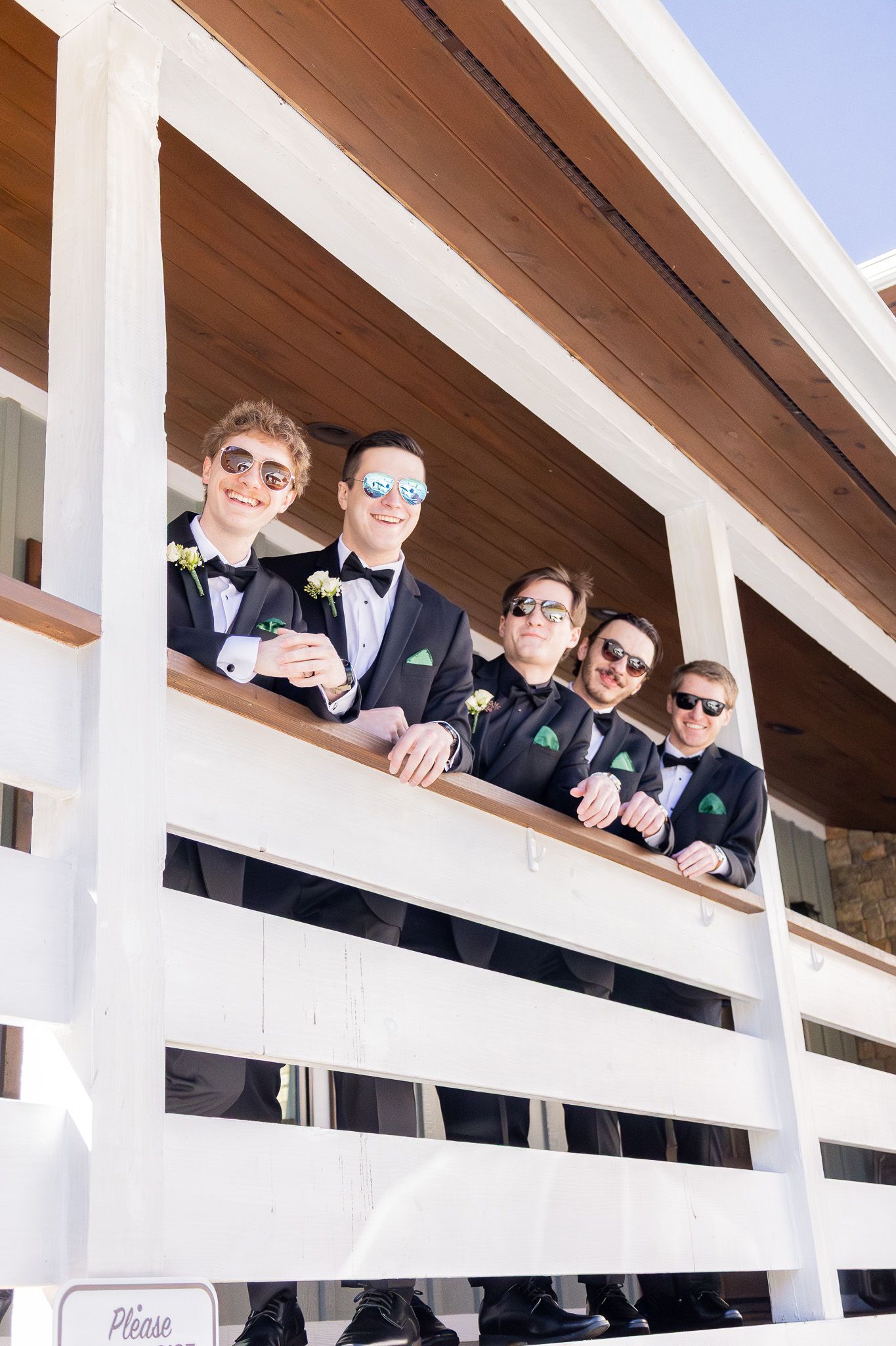 Five groomsmen in black tuxedos and sunglasses pose on a white wooden porch railing on a sunny wedding day.