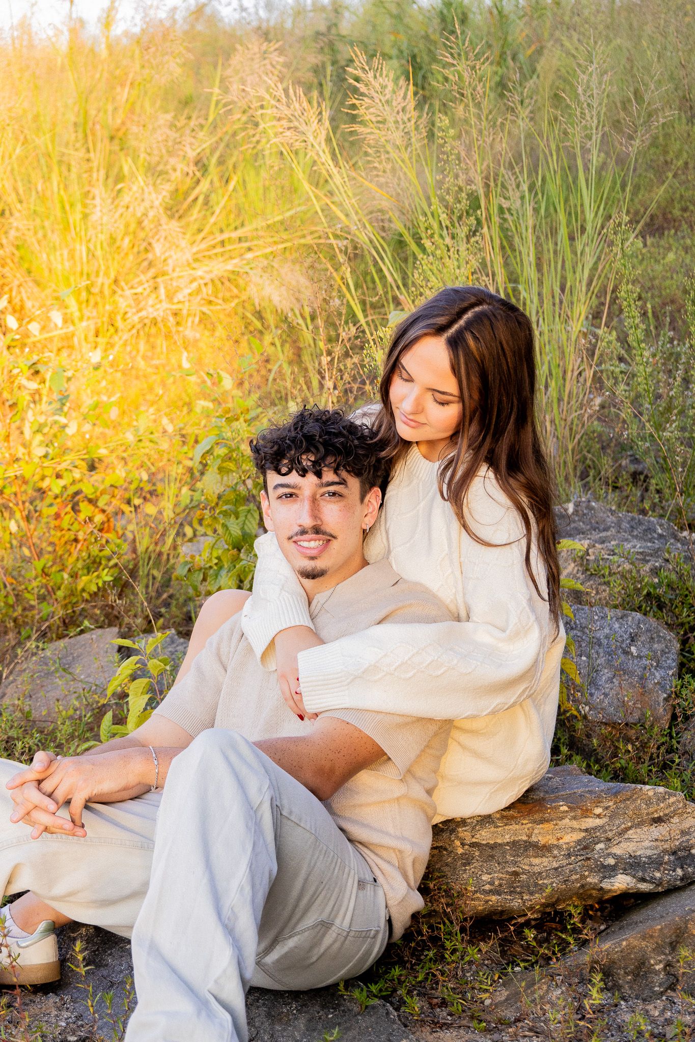 Couple sitting on rocks outdoors, woman embracing man from behind, surrounded by golden sunlit tall grasses.