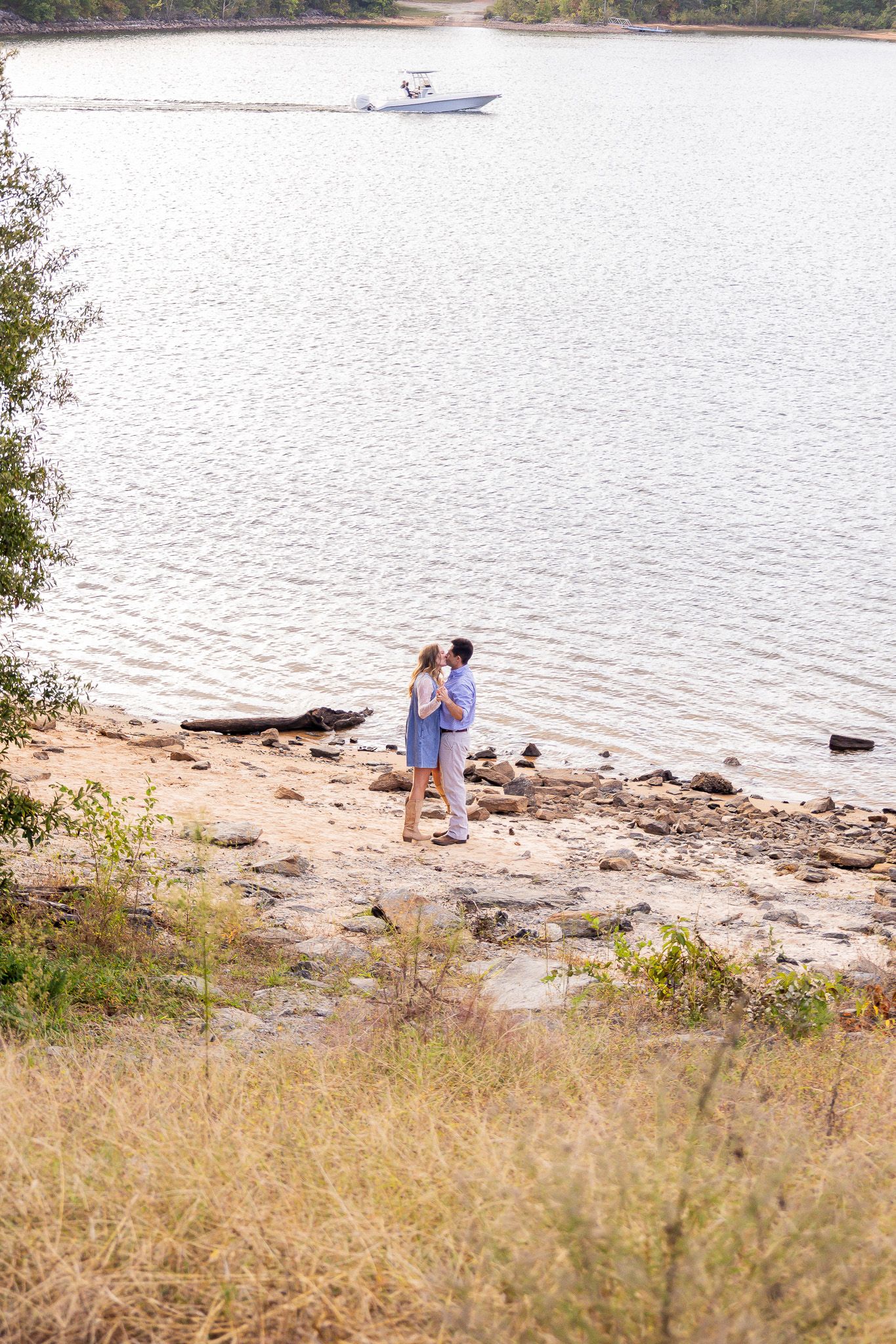 Couple embracing on a rocky beach shoreline with a motorboat passing on the calm lake behind them.