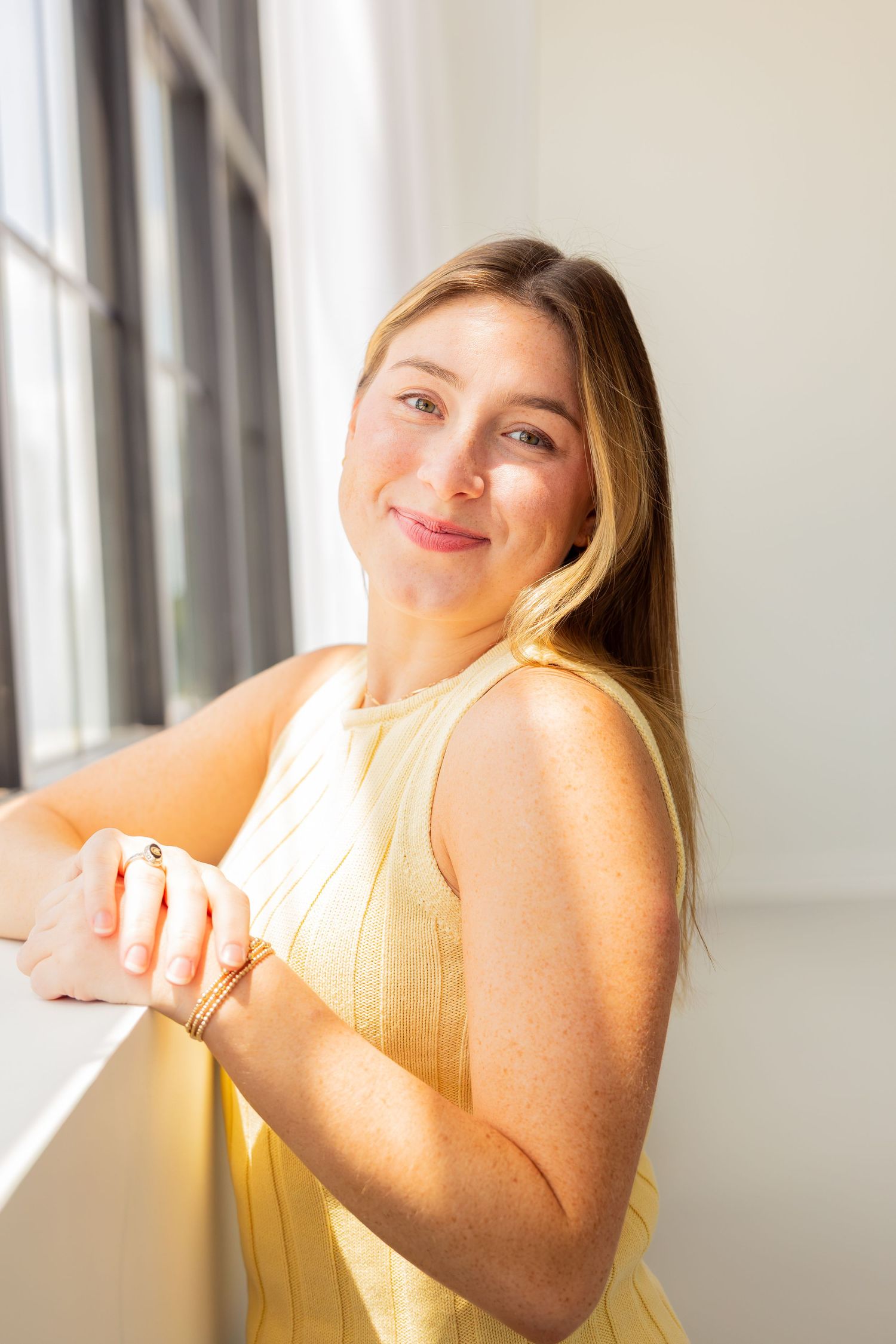 Young woman in yellow sleeveless dress smiling warmly by a sunlit window, wearing gold bracelets.