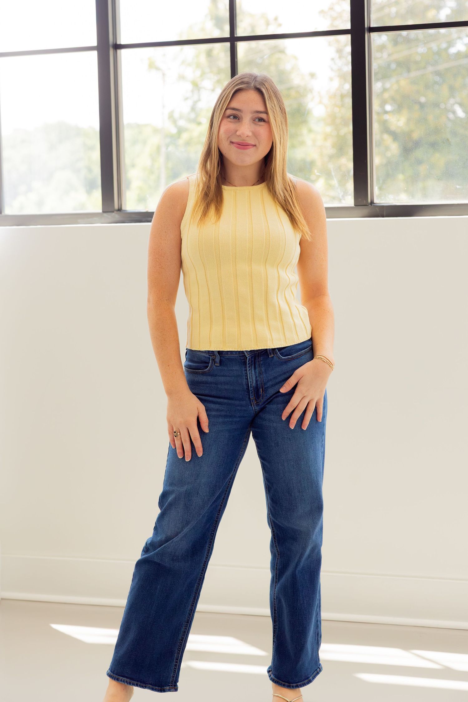 Woman wearing a yellow sleeveless ribbed knit top with wide-leg dark wash jeans, posing in a bright room.