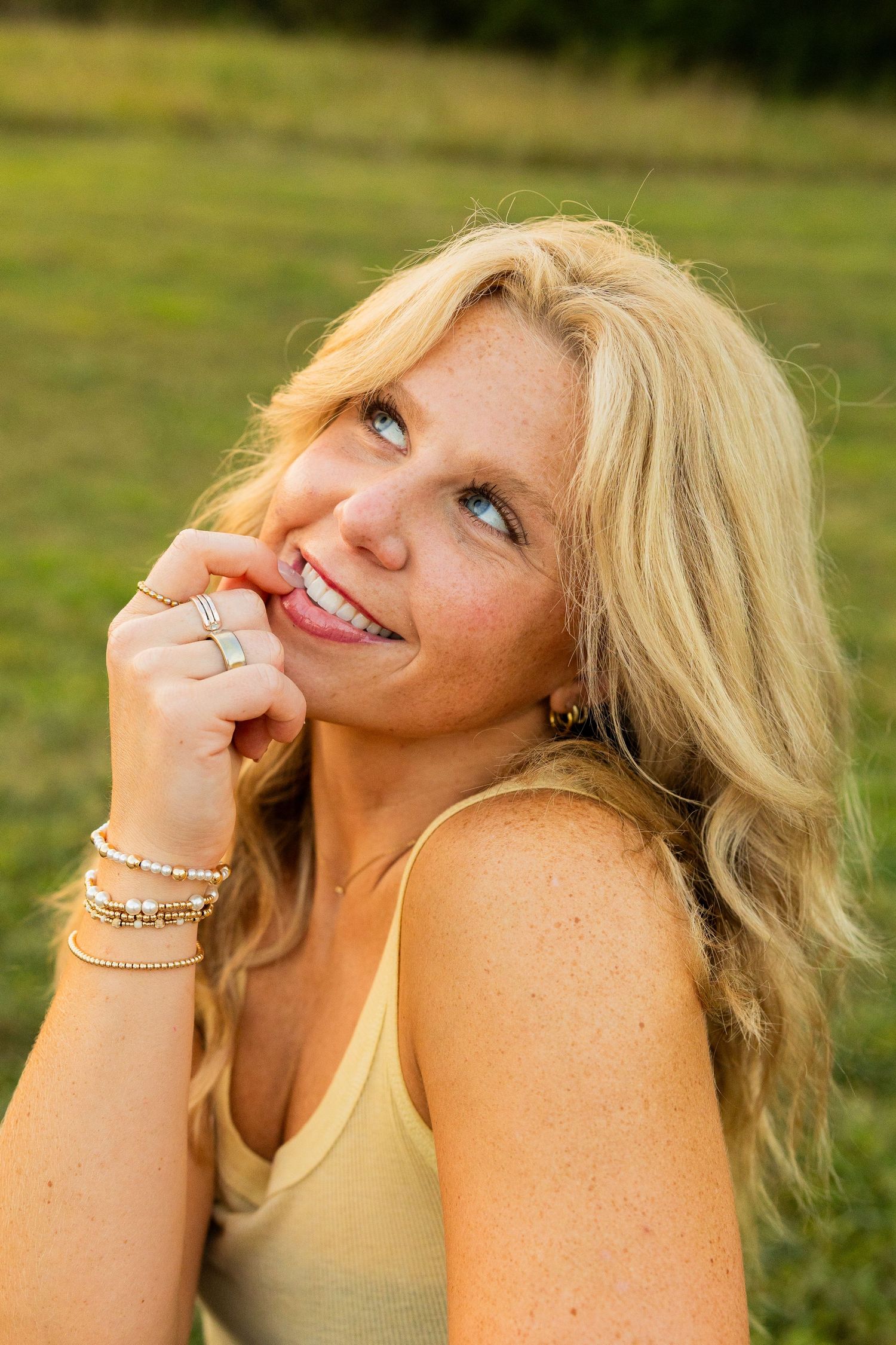 Smiling blond woman with freckles and blue eyes posing outdoors in a green field wearing gold jewelry.