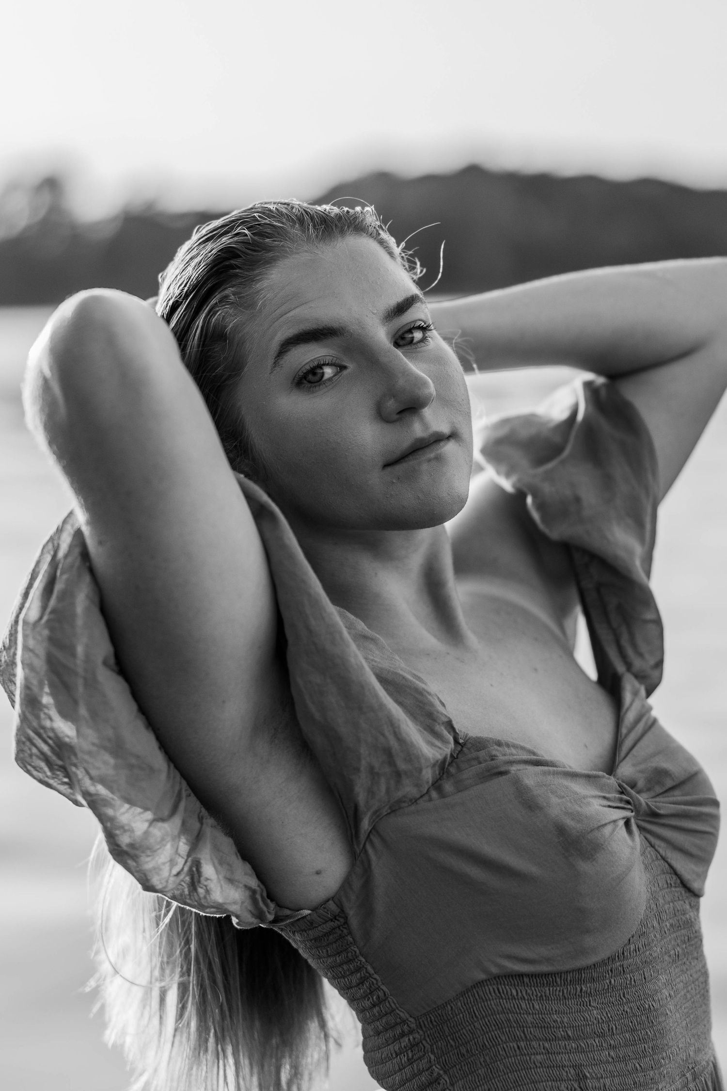 Black and white portrait of a young woman posing outdoors near water, hands behind head, wearing a tied crop top.