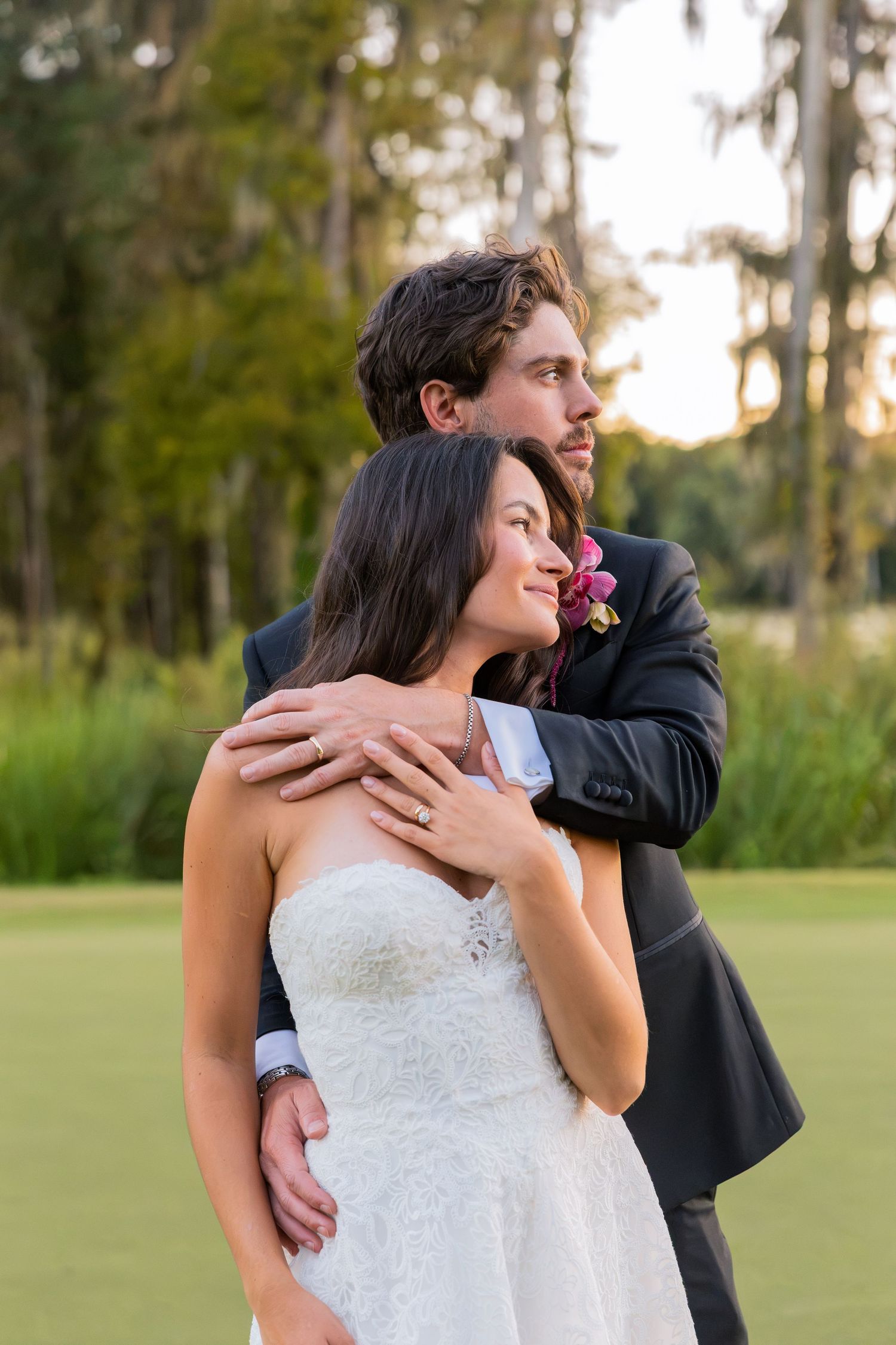 Bride and groom embrace outdoors at sunset, wearing wedding attire with lush green trees in the background.