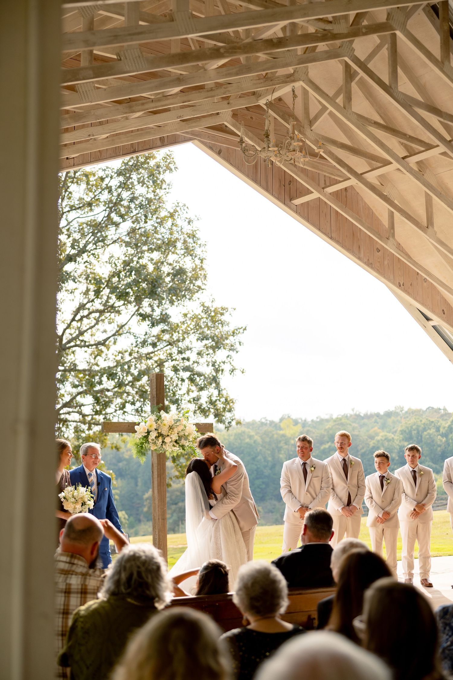 Outdoor barn wedding ceremony with bride and groom kissing at altar, groomsmen in tan suits, guests seated, lush green landscape backdrop.