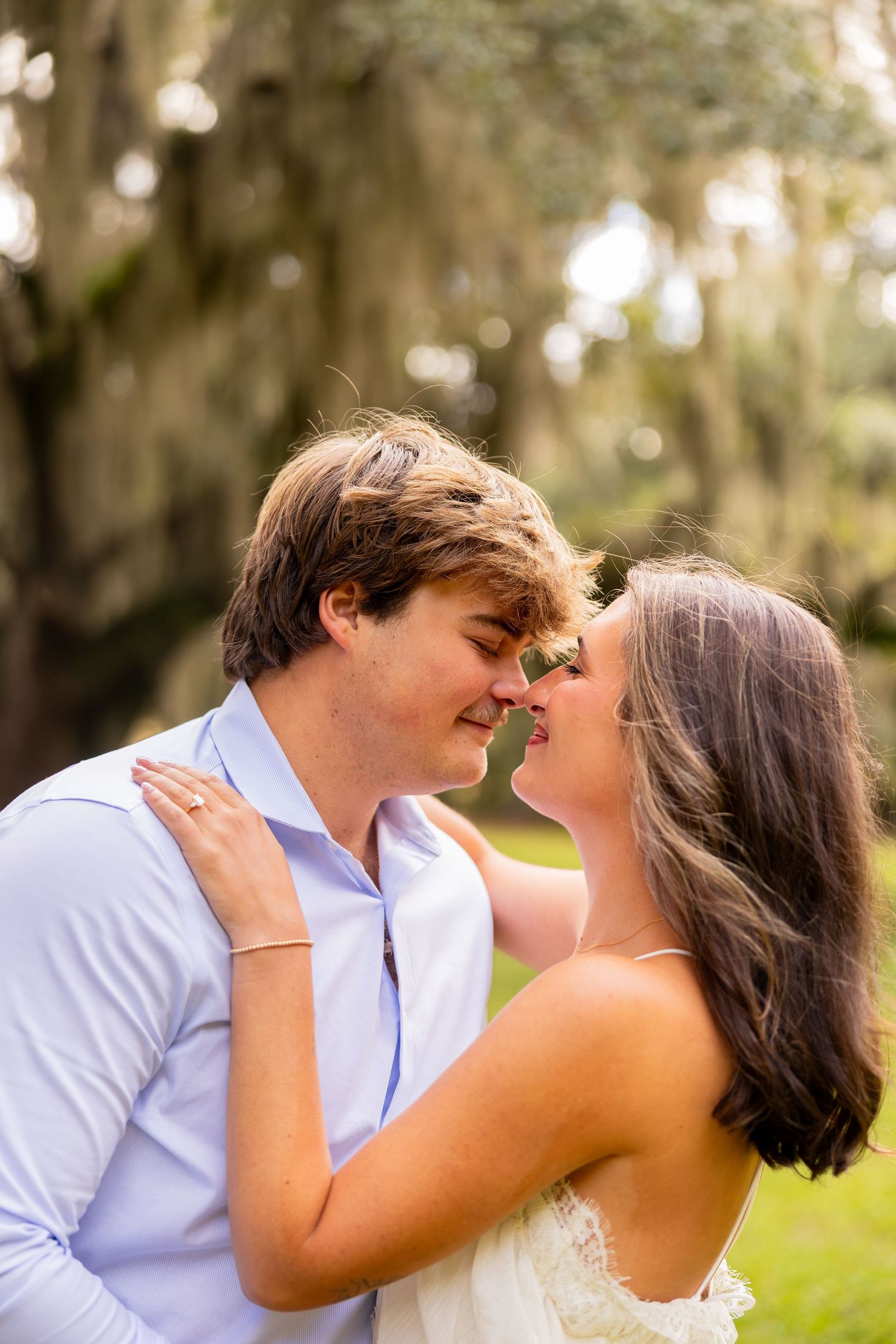 Engaged couple sharing an intimate moment outdoors, foreheads touching, under oak trees draped in Spanish moss, with her ring visible.