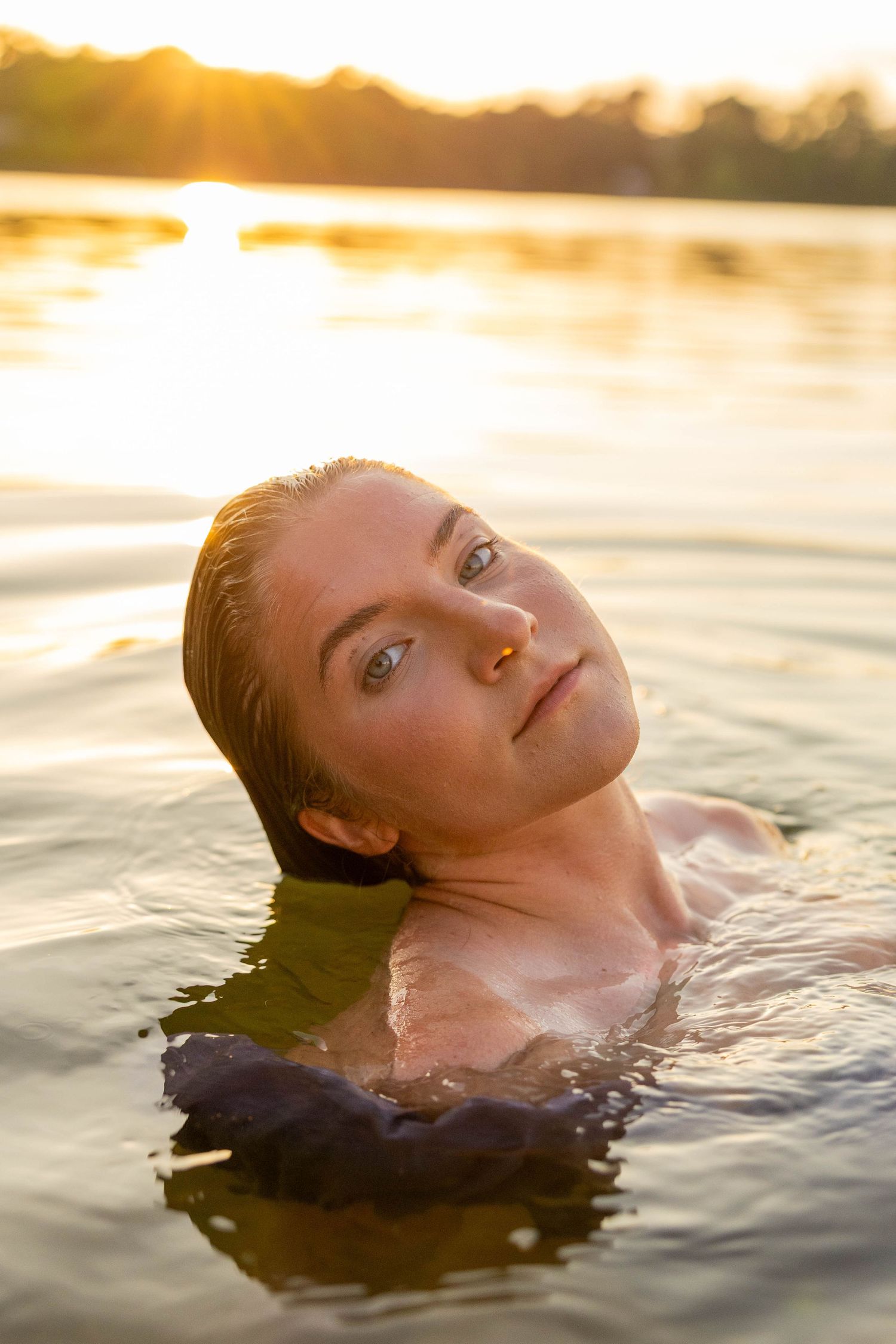 Woman with wet hair floating in glowing sunset-lit water, gazing upward peacefully.