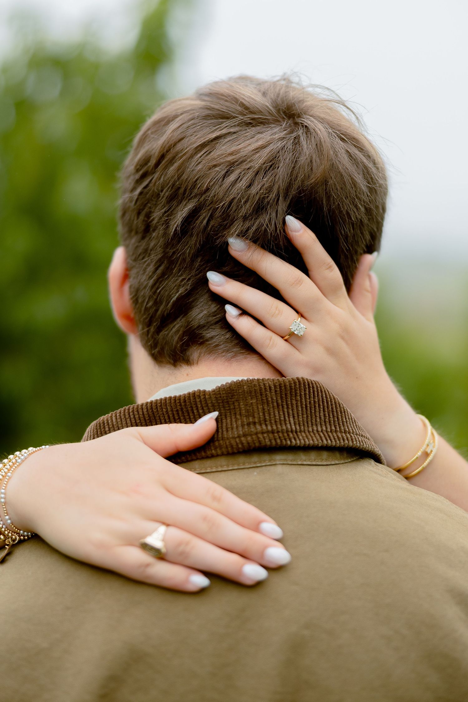 Woman with engagement ring and bracelet embracing man from behind, hands resting on his head and shoulder.