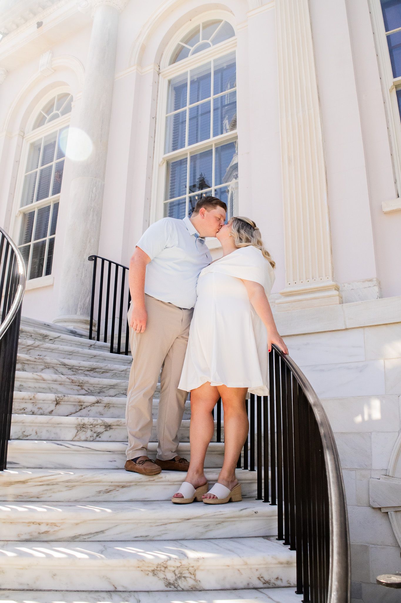 Couple sharing a romantic kiss on marble steps outside a grand white classical building with large arched windows.