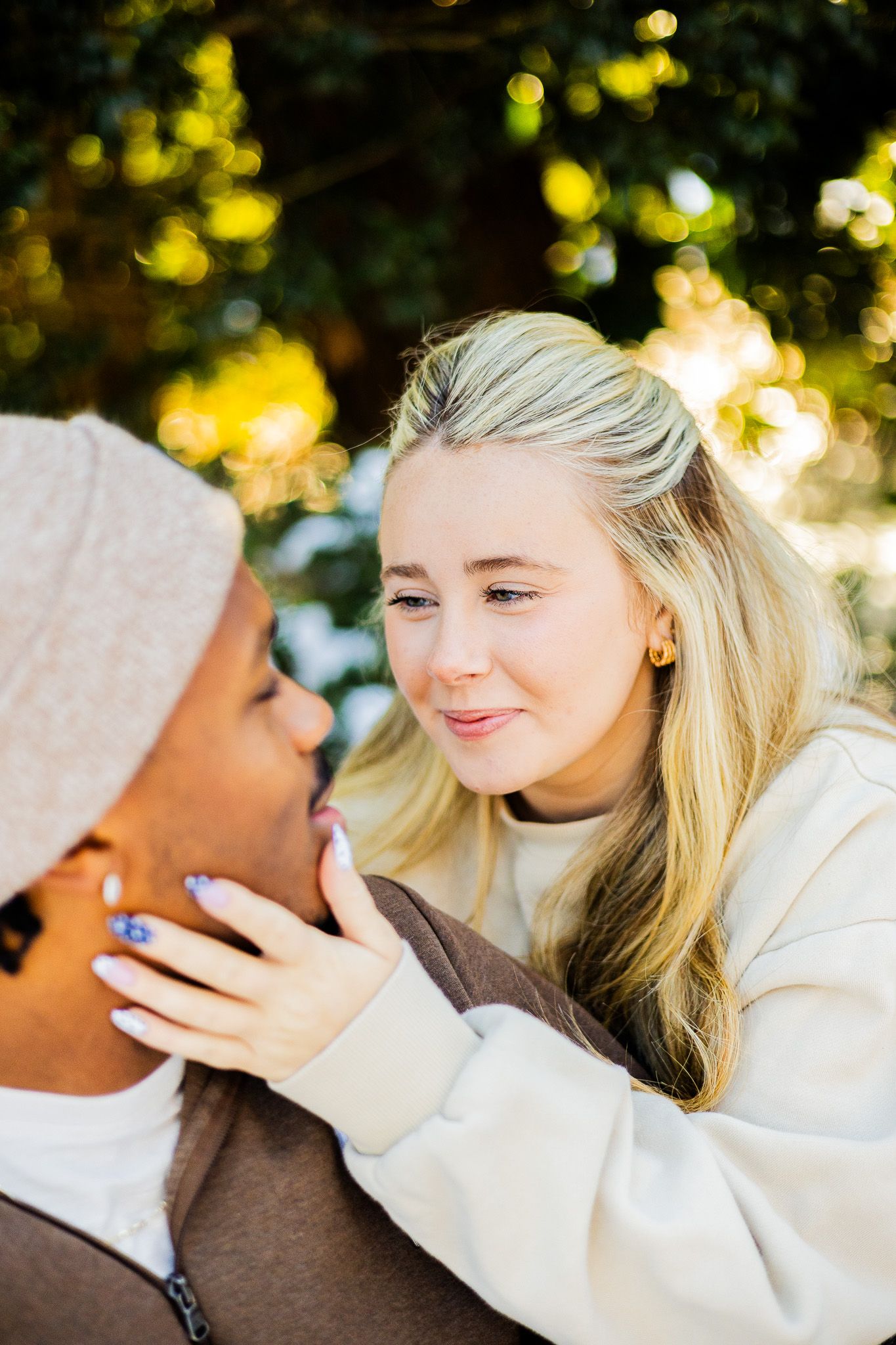 A blonde woman gently touches the face of her partner outdoors, sharing a tender moment with bokeh greenery background.