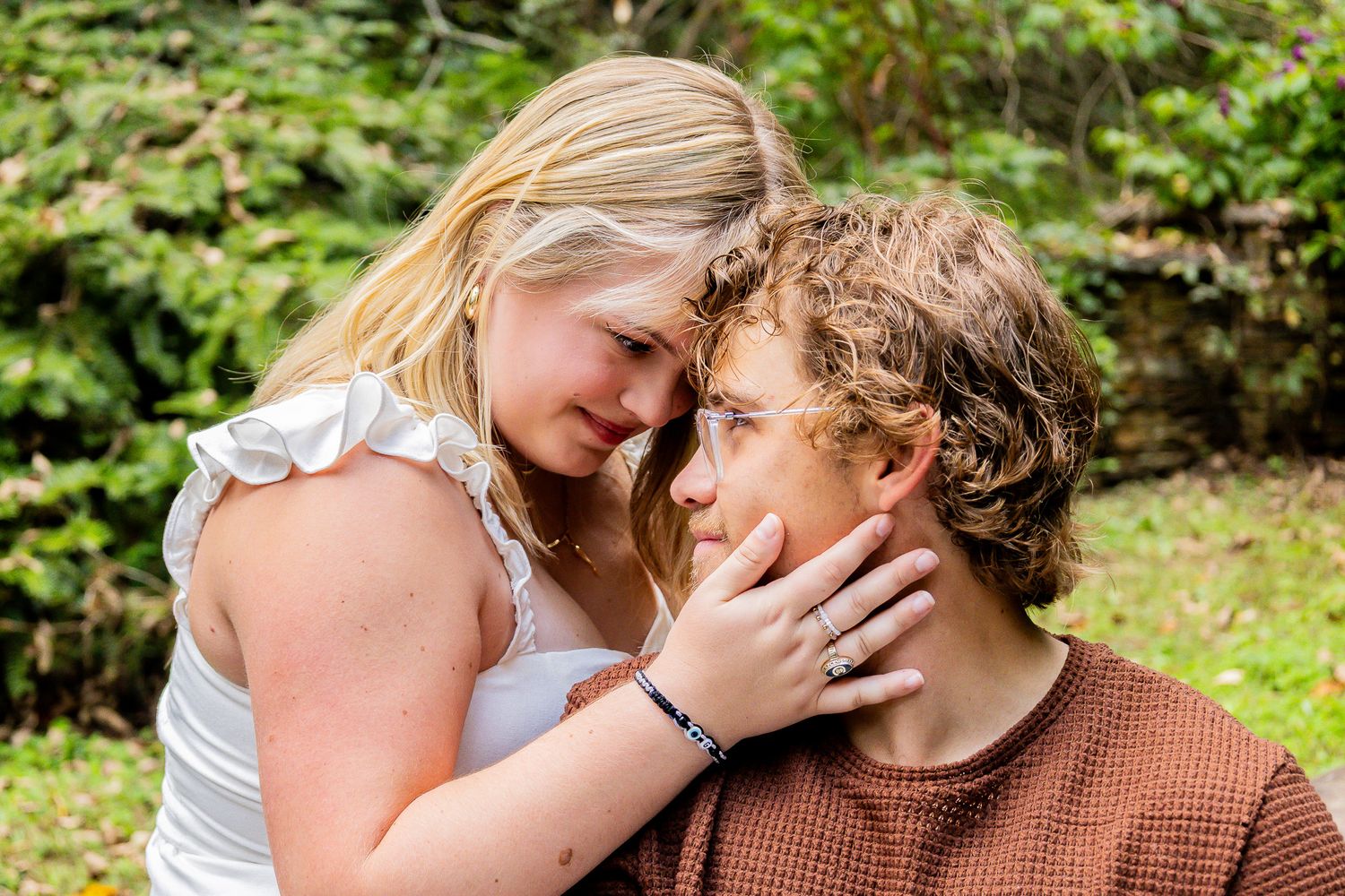 A young couple shares a tender romantic moment outdoors, with the woman gently touching the man's face near lush green foliage.
